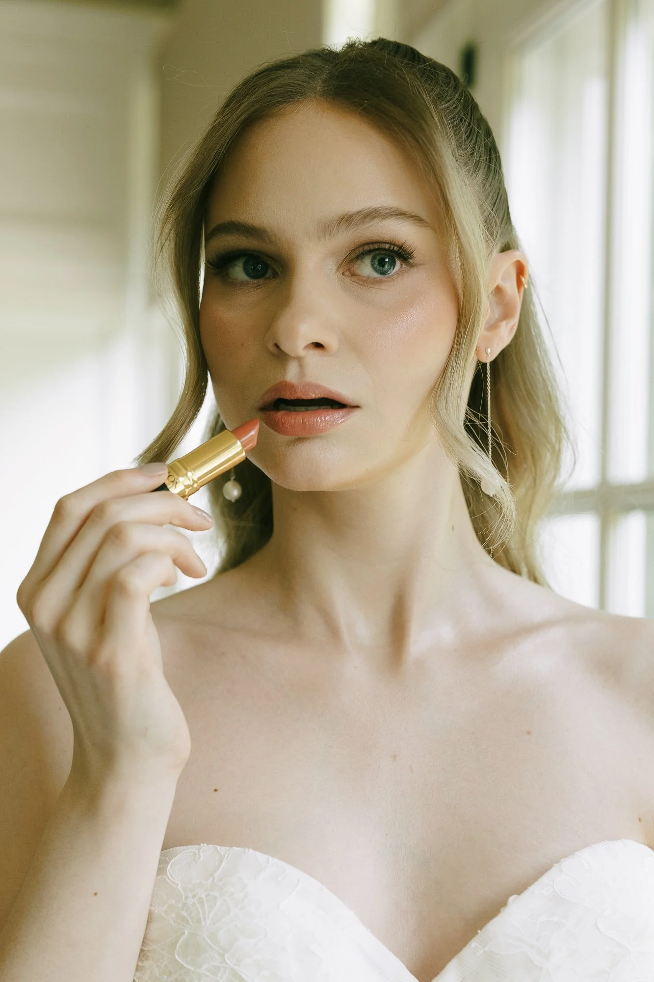 A woman with blonde hair applying nude lipstick, wearing a strapless white dress and long earrings, in front of a window.
