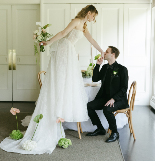 Bride in a white wedding dress holding a bouquet leaning over a seated groom in a black tuxedo, touching his forehead with her finger in a sunlit room.