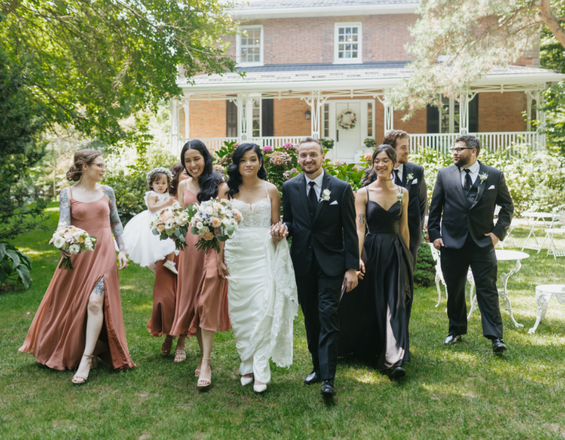 Group of wedding guests walking on green lawn in front of a brick house with white porch, celebrating a wedding.