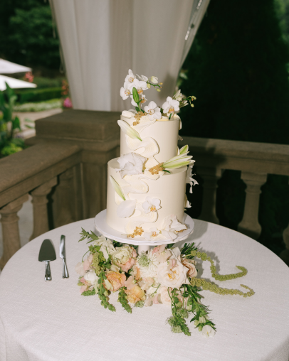 A three-tiered white wedding cake decorated with white flowers and green leaves, placed on a round table with a floral arrangement at the base.