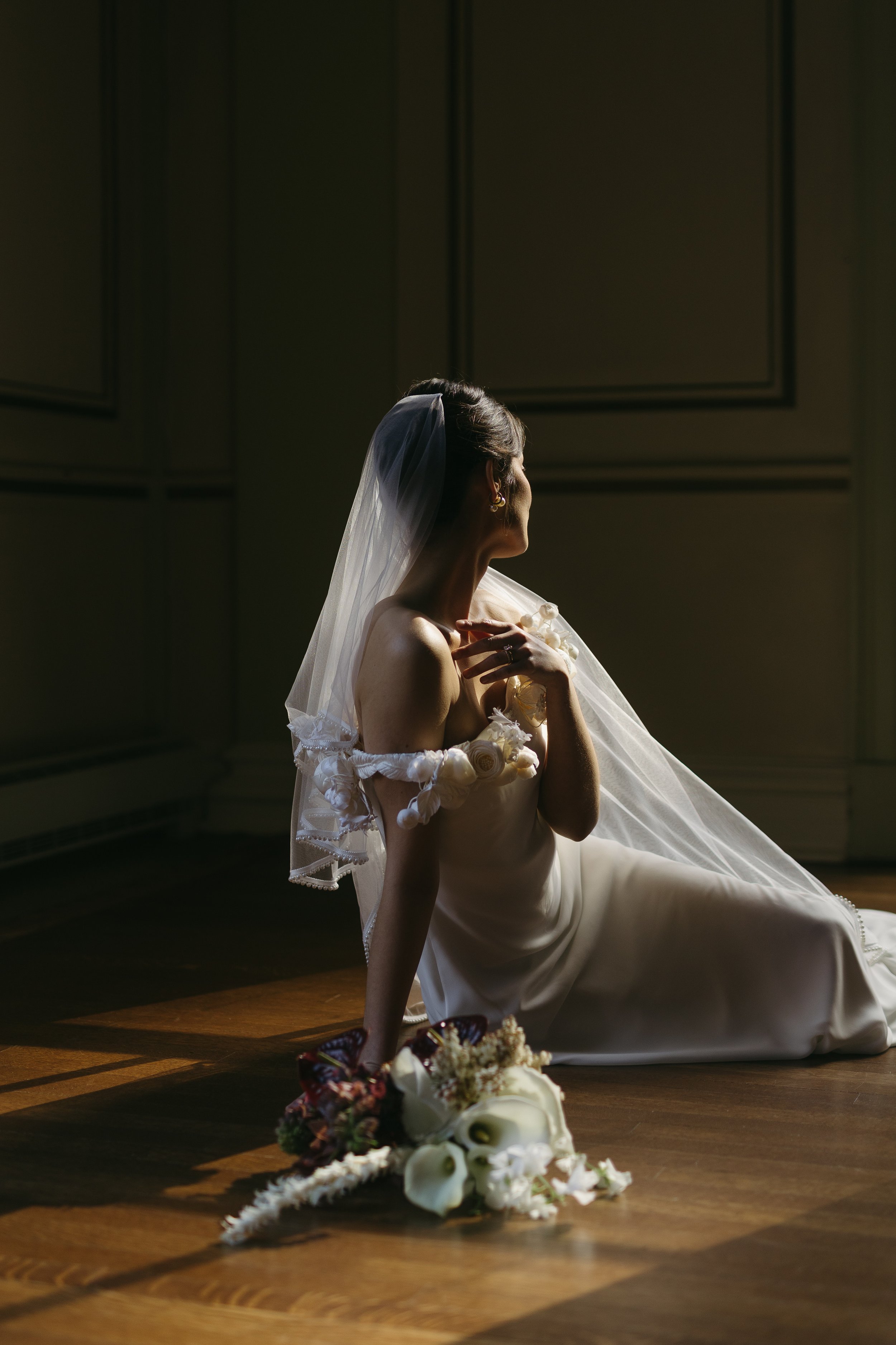 A bride sitting on the floor in a dimly lit room, with a veil on her head, wearing a strapless wedding gown, and surrounded by a bouquet of flowers.