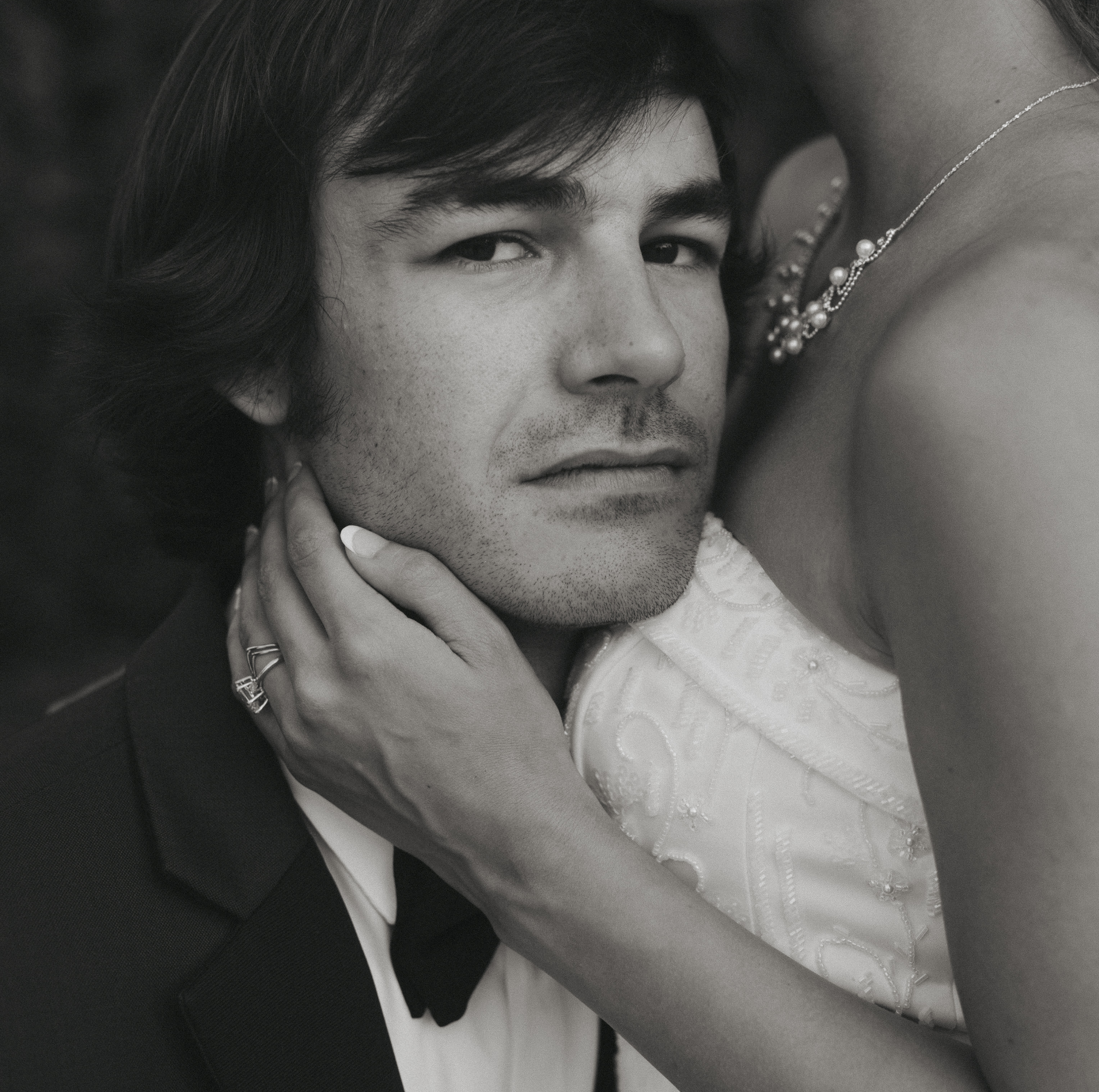 A black and white close-up photograph of a young man with dark hair and light skin, with his hand touching his neck, and part of a woman's arm and necklace visible in the frame.