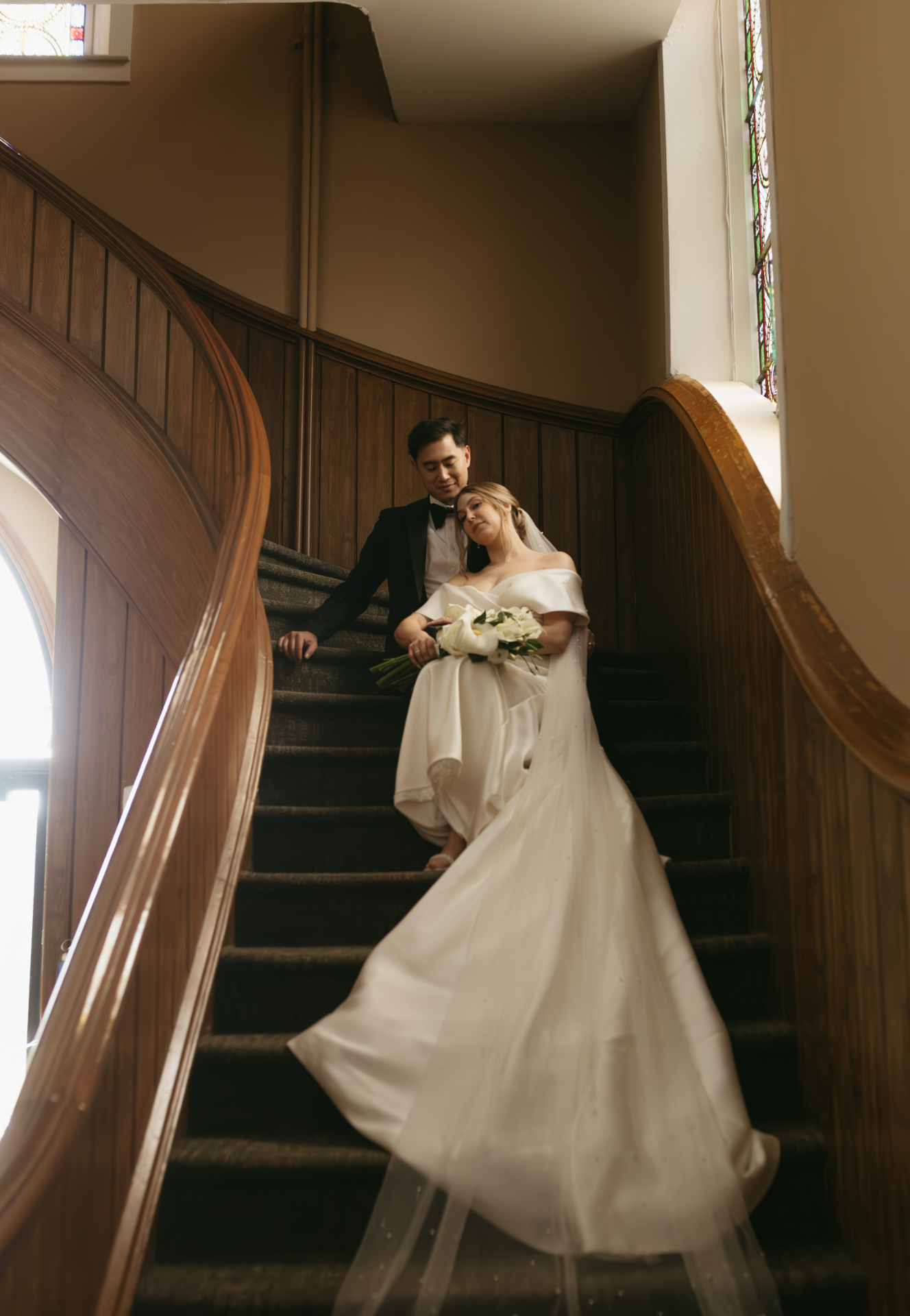 Bride and groom sitting on a staircase inside a church, with the bride holding a bouquet of white flowers, dressed in a white wedding gown, and the groom in a black tuxedo, leaning towards the bride.