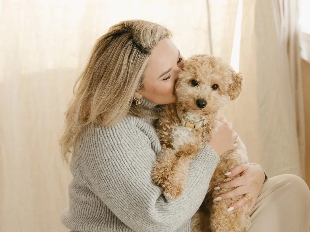 A woman with blonde hair in a gray sweater cuddles a small tan curly-haired puppy indoors.