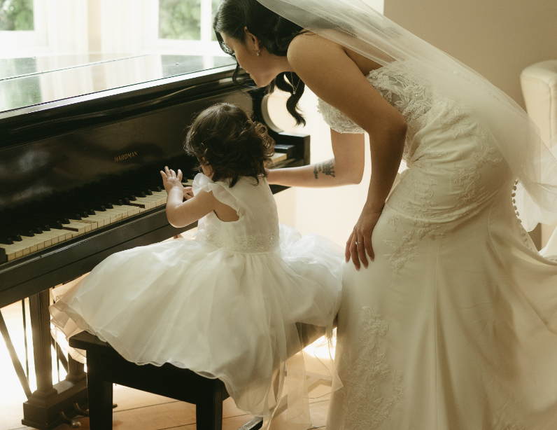 A bride helping a young girl play the piano in a sunlit room.