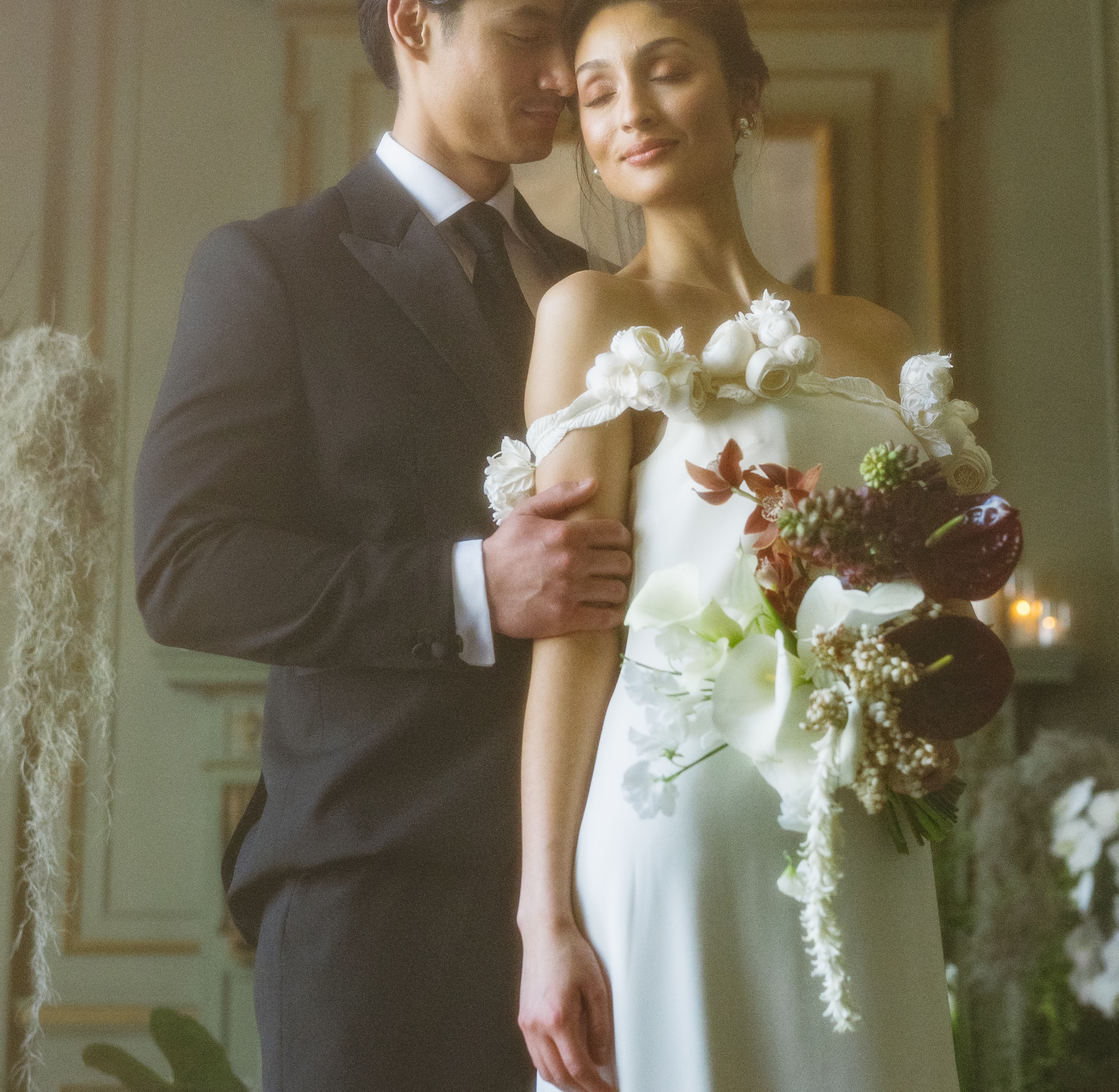 A bride and groom embracing, with the bride holding a floral bouquet, smiling softly with eyes closed.