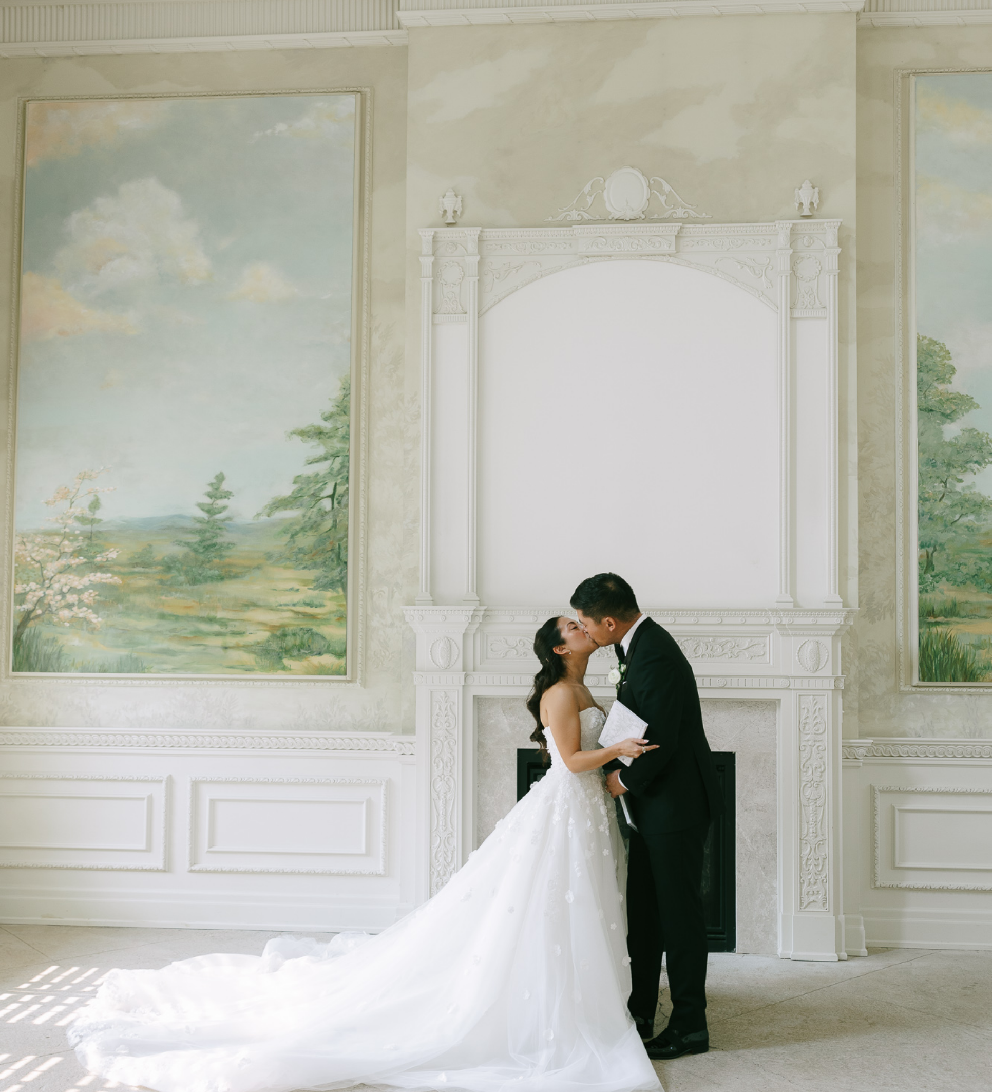 A bride and groom share a kiss during their wedding ceremony in a decorated room with landscape paintings, a white ornate fireplace, and a large white frame.