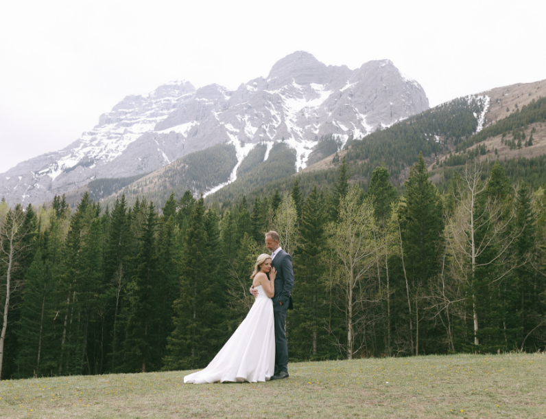 A bride and groom standing closely together in a field with a backdrop of dense green trees and snow-capped mountains.