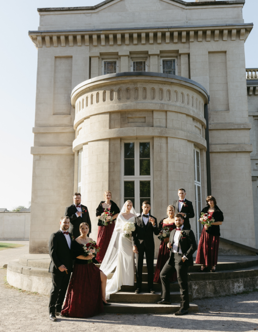 A bride and groom with their wedding party, standing on the steps in front of a large, historic building with rounded architecture and tall windows.