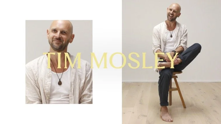 A split image of a bald man with a beard wearing a white shirt and black pants. On the left, he is shown from the chest up with the text "TIM MOSLEY" overlaid in large yellow letters. On the right, he is sitting on a wooden chair with his legs crossed, barefoot, against a plain white wall and light wooden floor, smiling and looking to the right.