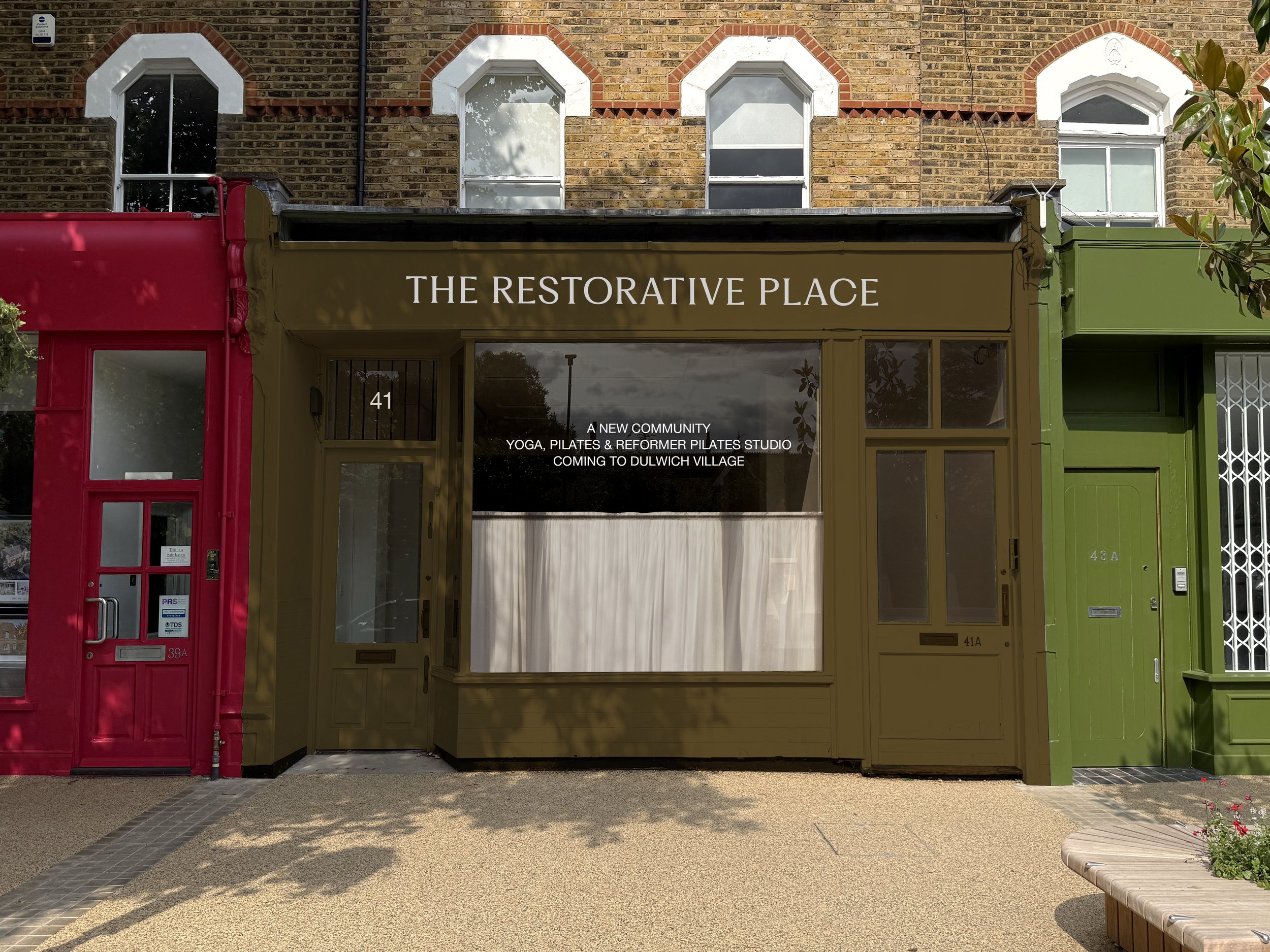 Storefront with sign 'The Restorative Place' offering yoga, Pilates, and reformer Pilates, located at 41 Dulwich Village, with a red door on the left and a green door on the right, brick building in the background.