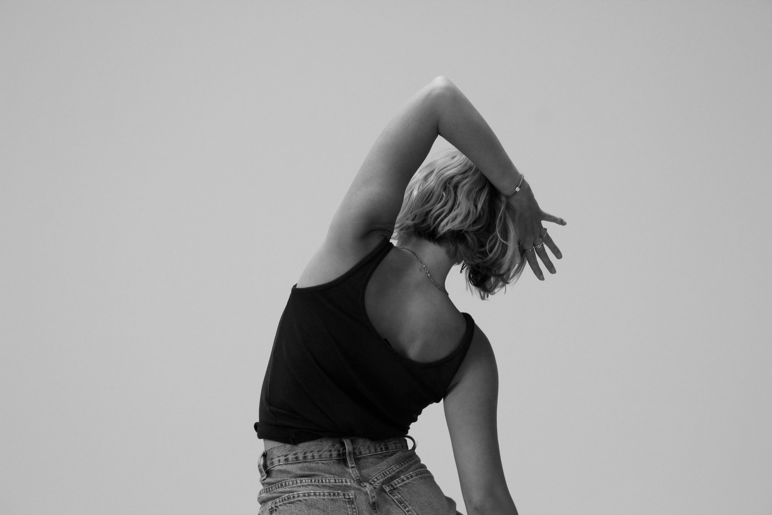 Black and white photo of a woman stretching with her head tilted back, fist pointing upward, and arm bent over her head, wearing a tank top and jeans against a plain background.