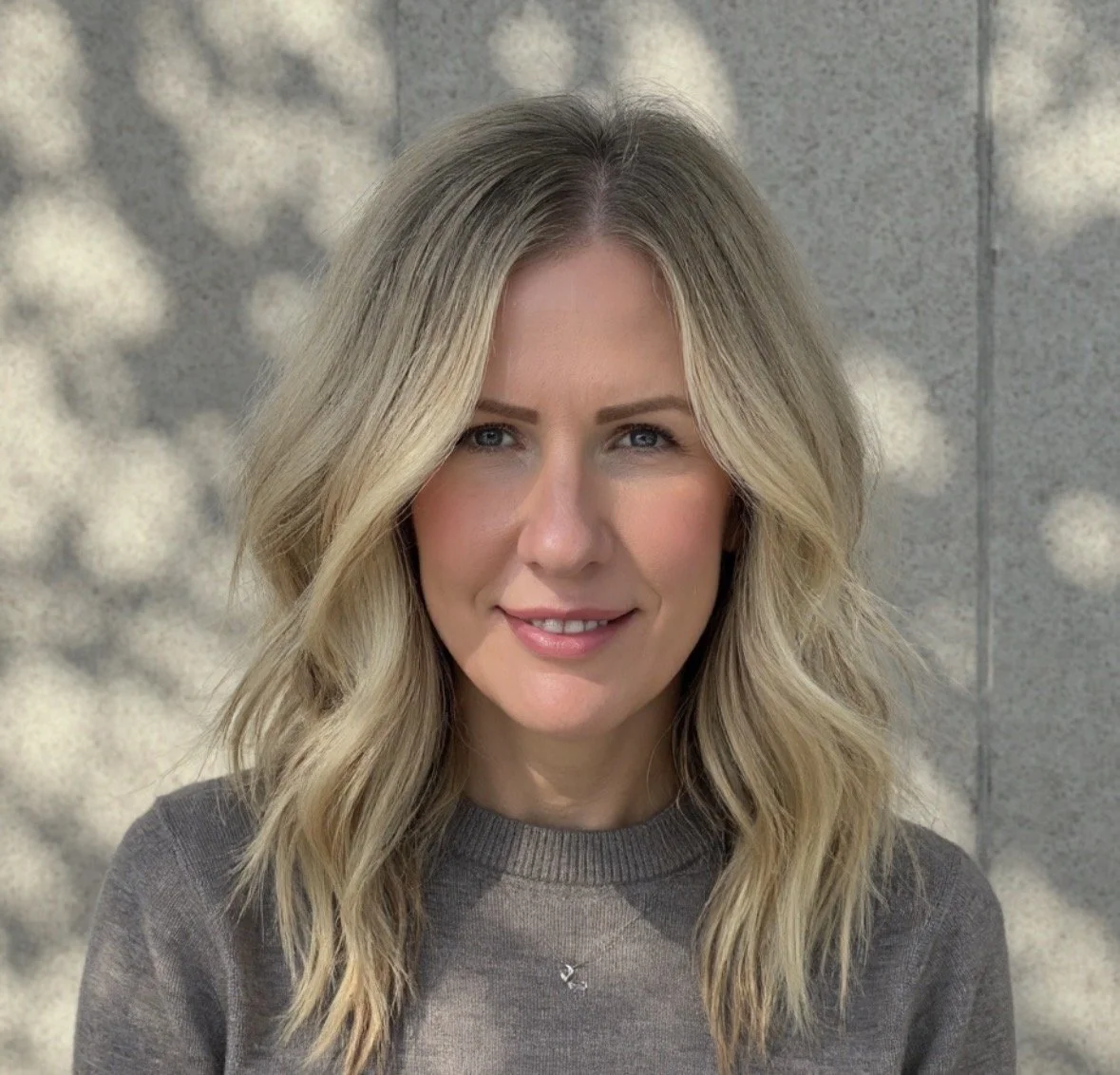 A woman with blonde wavy hair, wearing a gray sweater, standing in front of a textured wall with blurred shadow patterns.