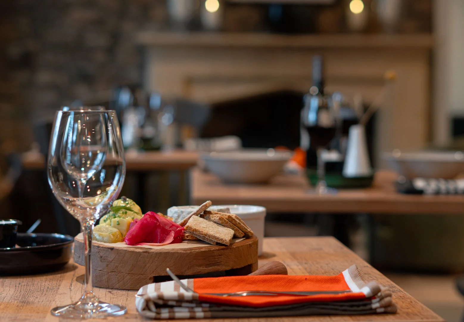 A restaurant table set with a wine glass, colorful appetizer with cheese, vegetables, and bread on a wooden serving board, a stack of plates, and an orange-striped napkin with utensils, in a cozy, rustic dining area.