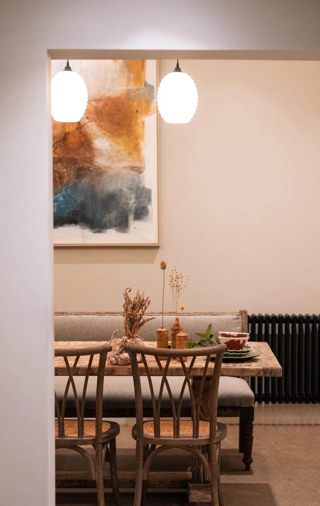 A dining room with a wooden table, four wooden chairs, and a gray upholstered bench. The table is decorated with dried flowers in vases and a green plant. Two hanging pendant lights and an abstract art painting are visible in the background.