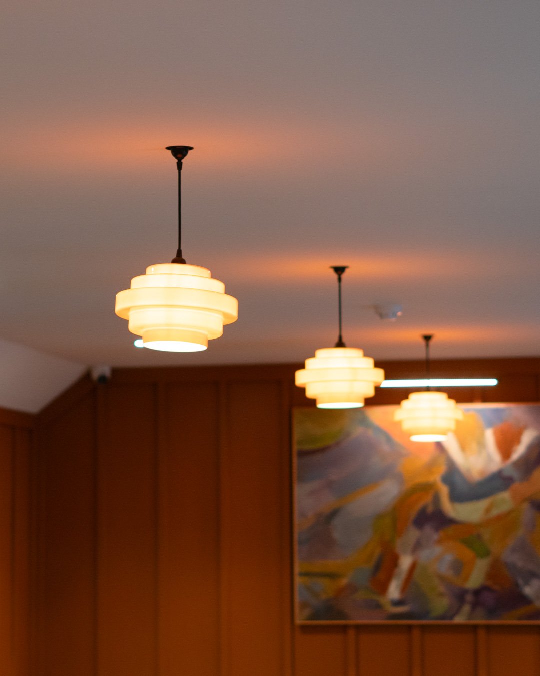 Interior view of a room with wooden panel walls, hanging ceiling lights, and an abstract painting on the wall.