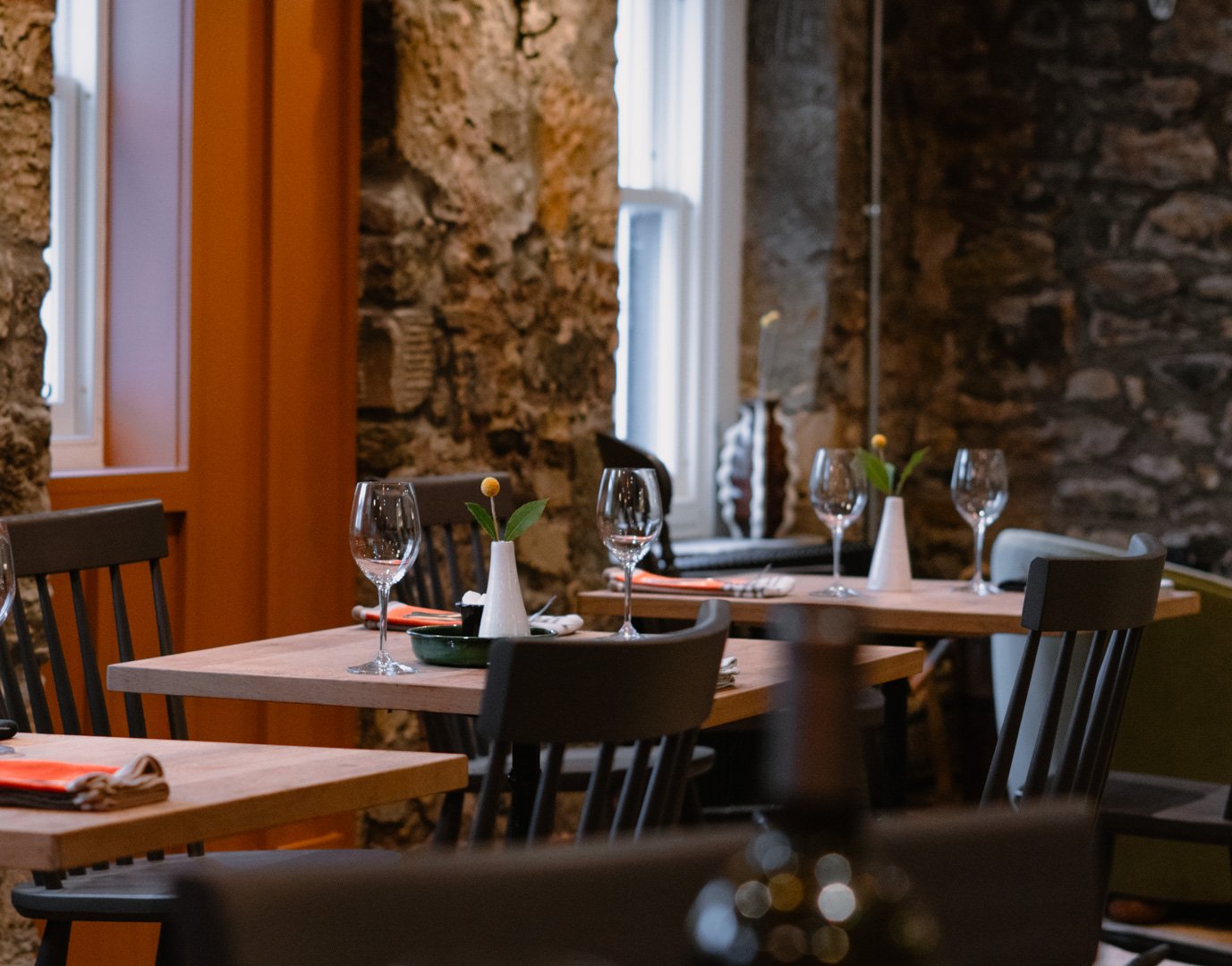 A cozy dining area with wooden tables, black chairs, and stone brick walls. The tables are set with wine glasses, small vases with yellow flowers and green leaves, and napkins with utensils. Natural light comes through the windows in the background.