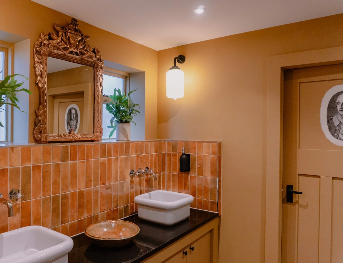 Bathroom with orange tiles, two white sinks, a decorative ceramic bowl, greenery on a window sill, a mirror with an ornate wooden frame, a wall-mounted light fixture, a black soap dispenser, and a door with a historical portrait.