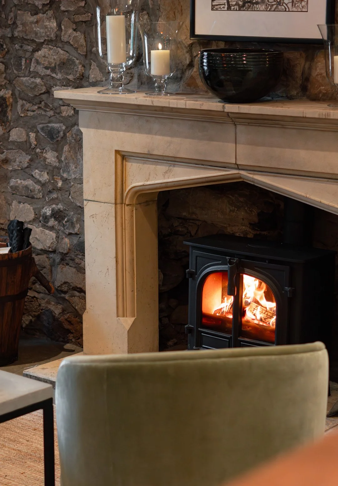 A cozy living room corner with a stone fireplace, a wood stove with a fire inside, a beige mantel decorated with candles in glass holders and a black bowl, and a framed wall art above the mantel.