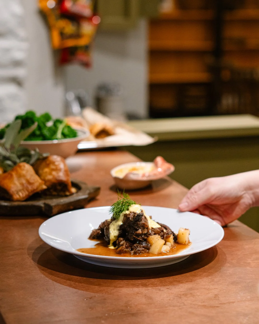 A plate of beef stew garnished with a sprig of dill, served with mashed potatoes, on a wooden table in a restaurant setting.