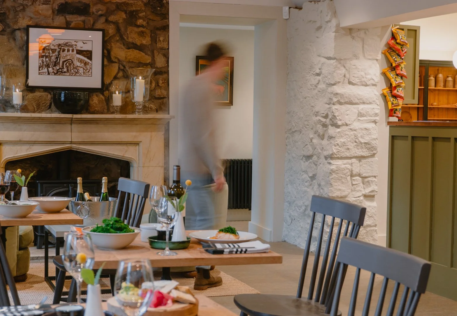 A dining room with a stone fireplace, a wooden table set with plates, glasses, and green napkins, with a person in motion blurred in the background near the doorway, and a corner with snack bags on the wall.
