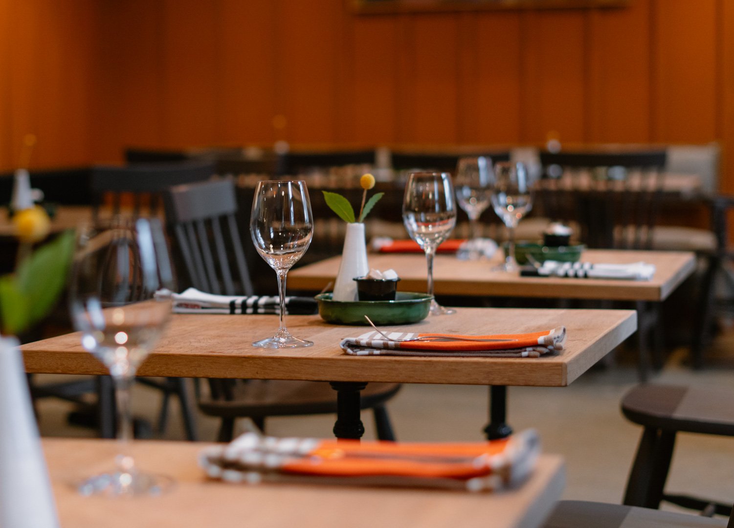 Restaurant dining table set with wine glasses, placemats, and a small vase with a flower, in a warmly lit dining room.