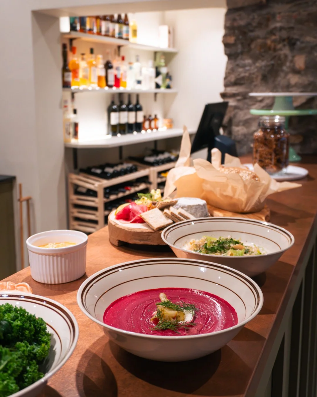 Bowls of colorful soups and plates of bread and vegetables on a wooden counter in a cozy restaurant with bottles on shelves in the background.