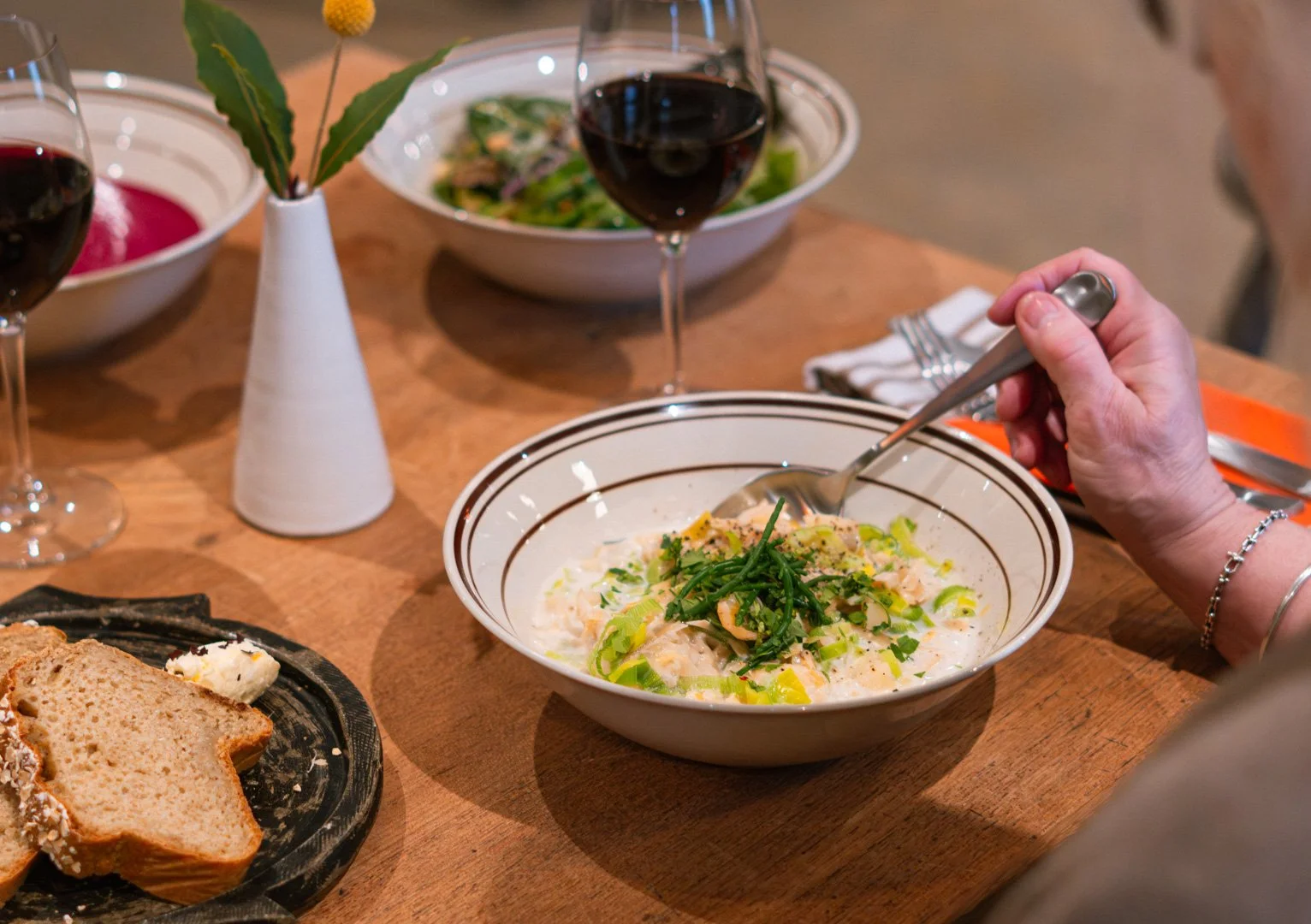 A person eating a bowl of creamy seafood chowder with green onions garnished on top, at a dinner table with glasses of red wine, salad, bread slices, and a vase with yellow flowers.