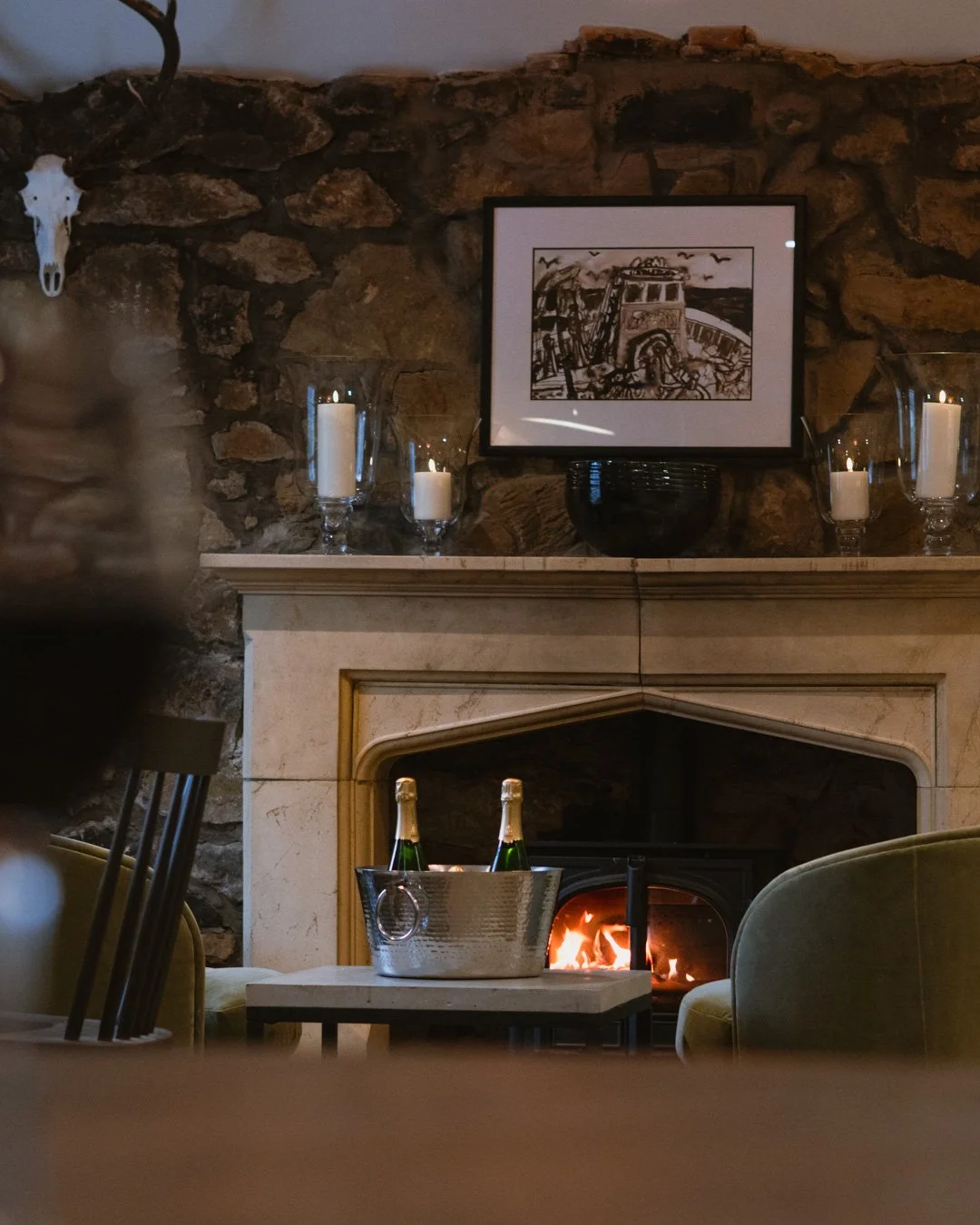 A cozy living room with a stone fireplace surrounded by candles and a black bowl on the mantel. A framed black and white drawing is hung above the mantel. In front of the fireplace, there is a small table with a silver bucket holding champagne bottles, and an armchair is partially visible on the right.