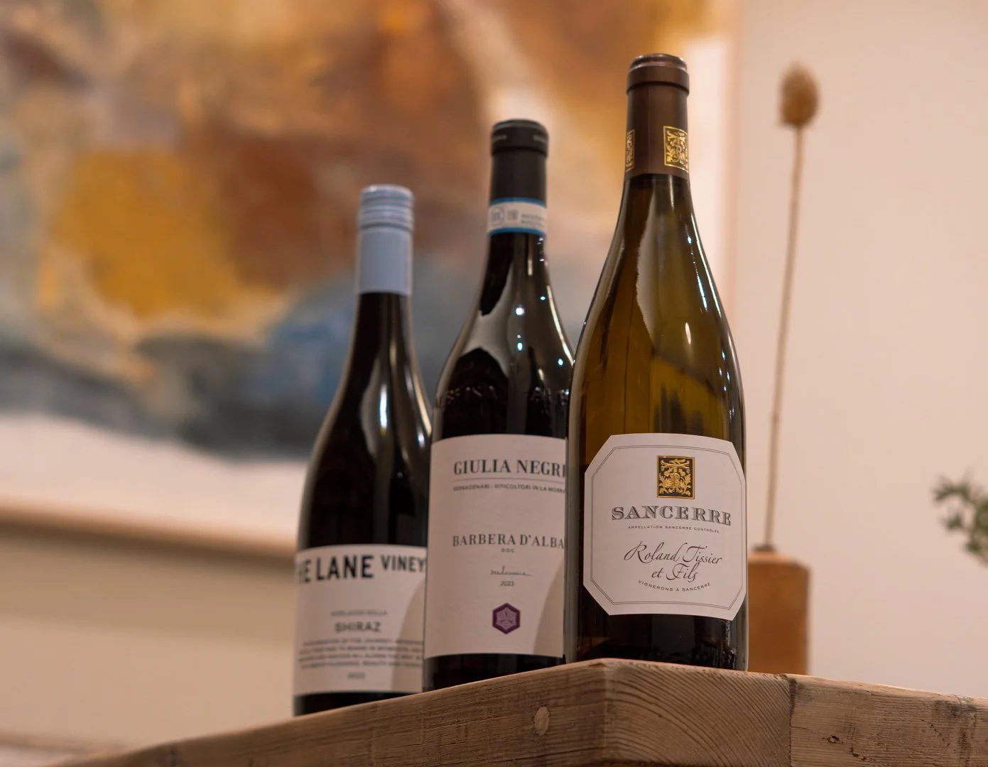 Three bottles of wine placed on a wooden surface with a blurred abstract painting in the background.