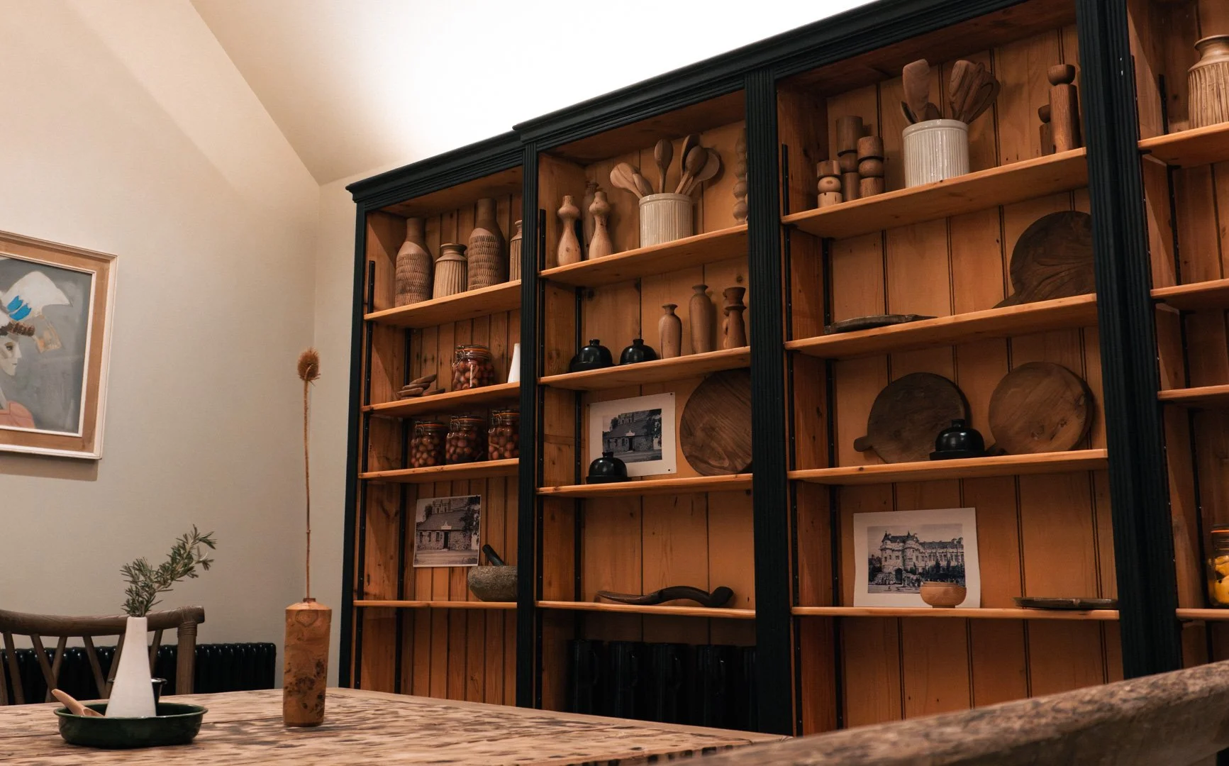 Interior of a room with a large wooden shelf displaying various wooden bottles, bowls, and jars, next to framed photographs, with a small table holding a plant and a ceramic vase.