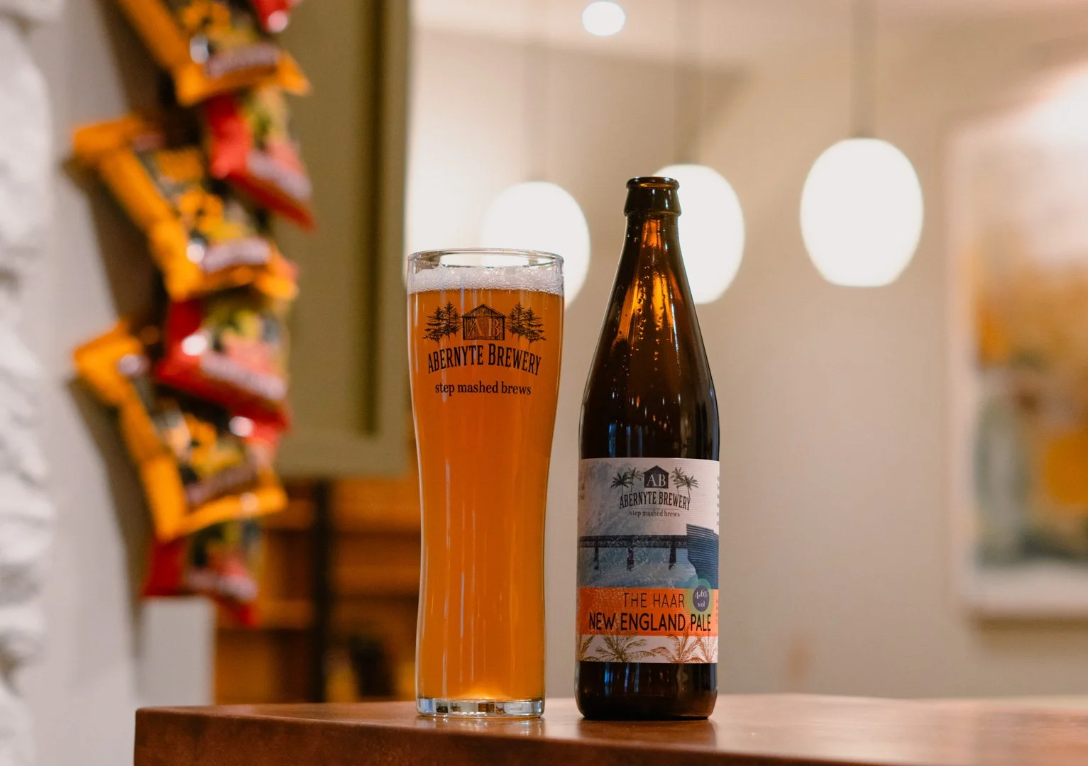 A glass of beer and a bottle of beer standing on a wooden surface inside a brewery or bar. The glass has the logo and text for Abernyle Brewery and the bottle has a label for The Haar New England Pale.