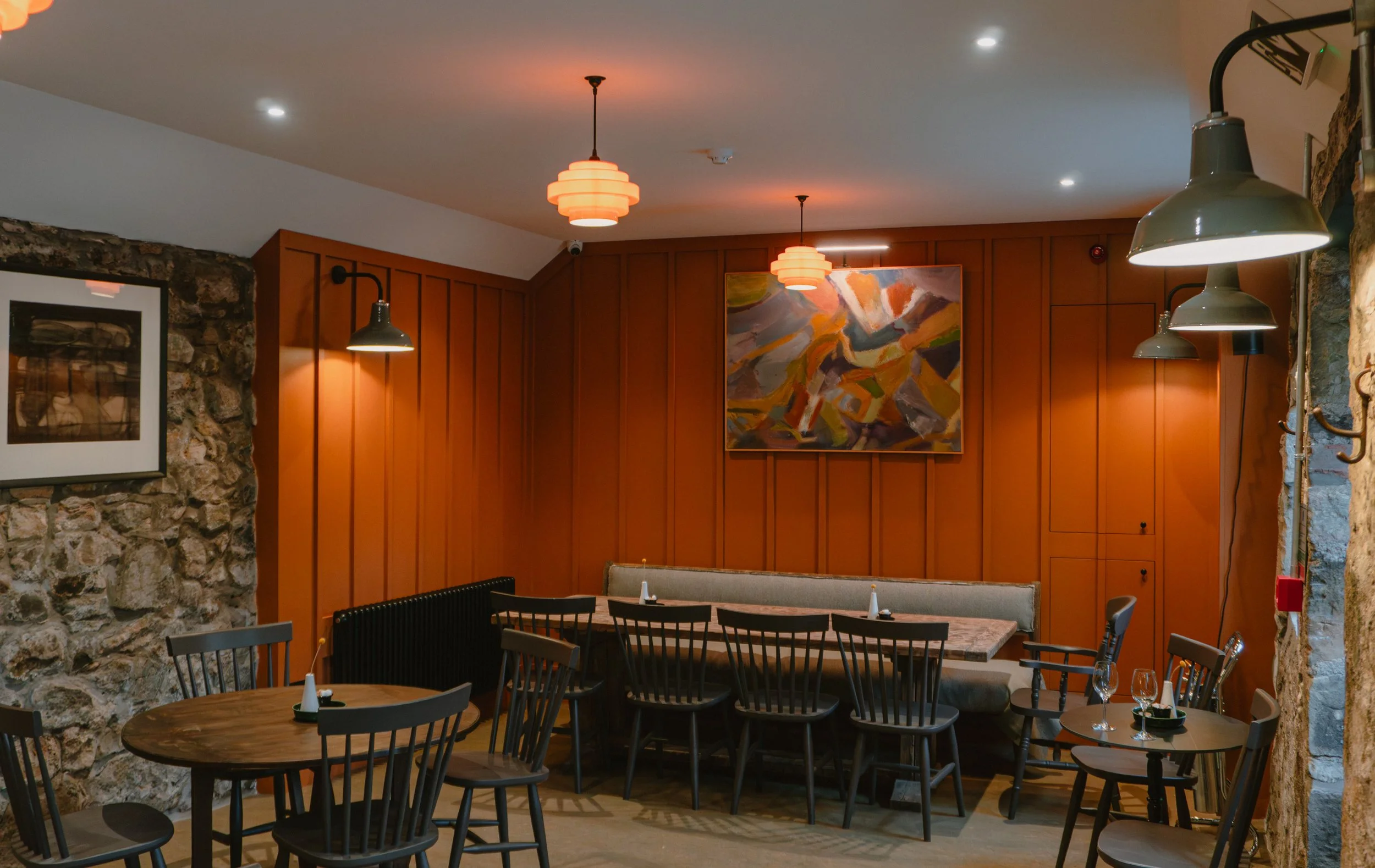 Interior of a cozy restaurant with wooden walls, stone accents, framed artwork, pendant lights, tables set with glasses and napkins, and comfortable chairs.