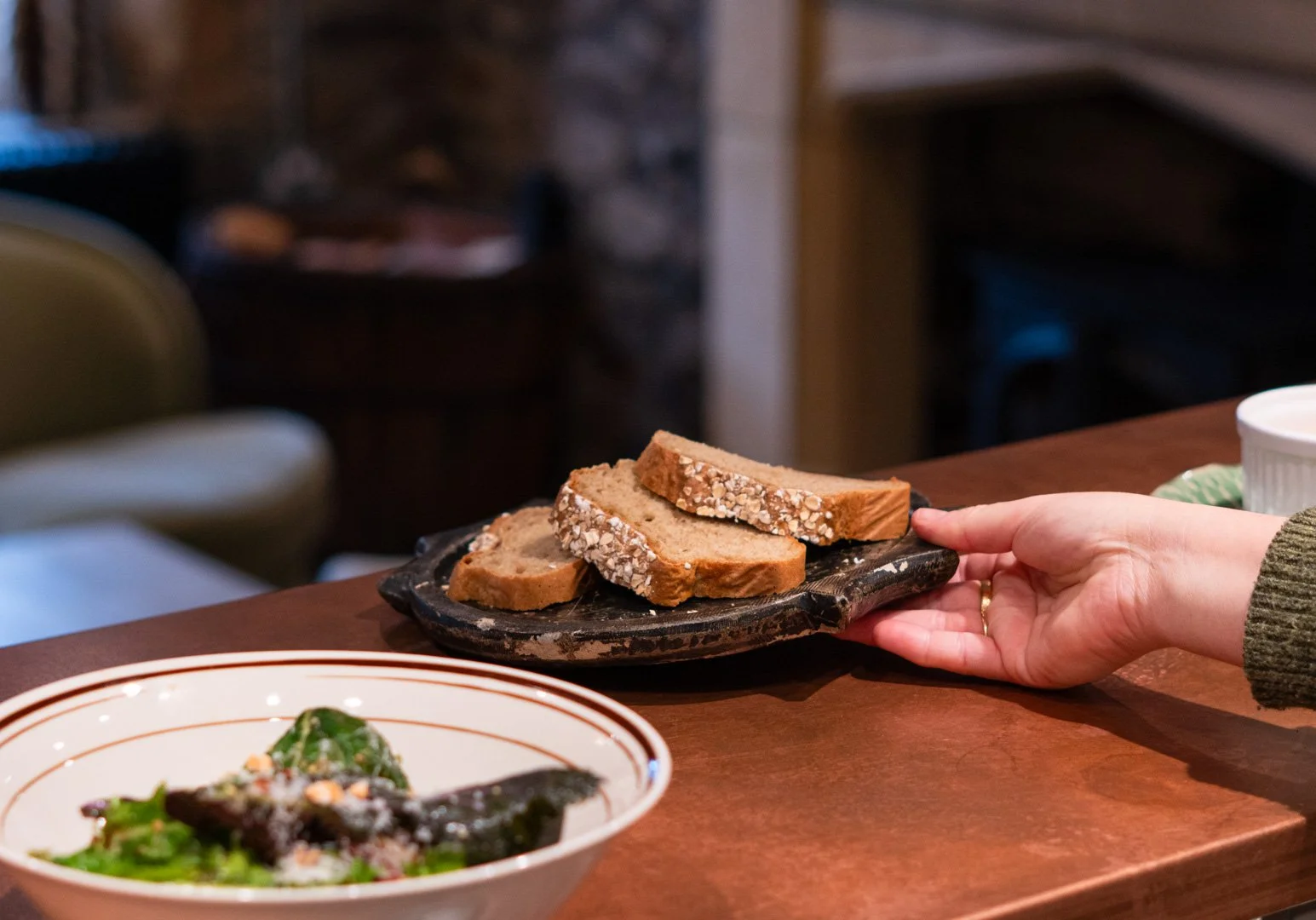 A person with a ring on their finger is holding a black tray with three slices of bread with oats on the crust. In the foreground, there is a bowl with greens and what appears to be a piece of cooked meat, possibly fish.