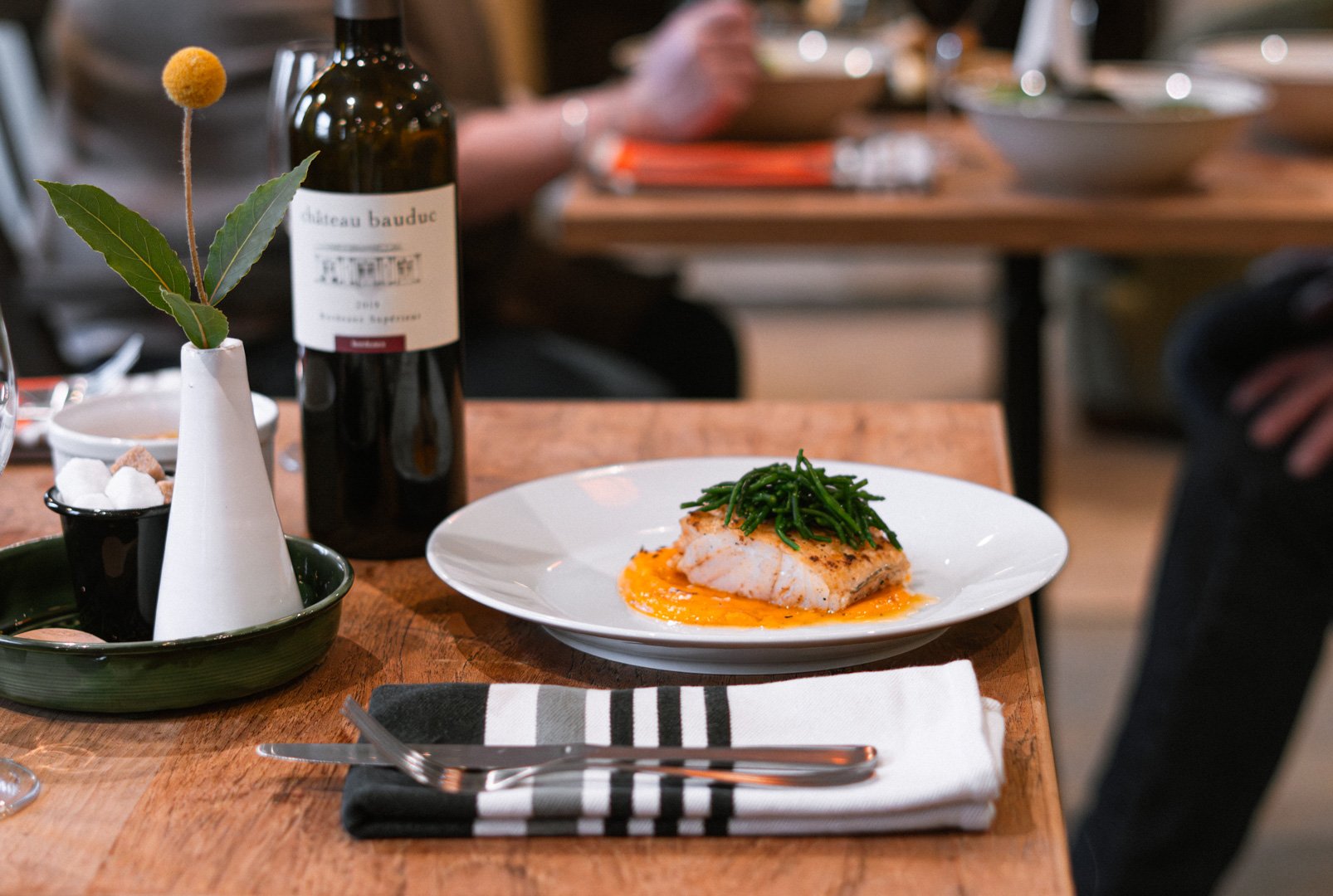 A plated serving of cooked white fish topped with green beans, on an orange sauce on a white plate on a wooden table. There is a bottle of red wine, a small vase with leaves and an orange flower, salt and pepper shakers, and a napkin with a fork and knife.