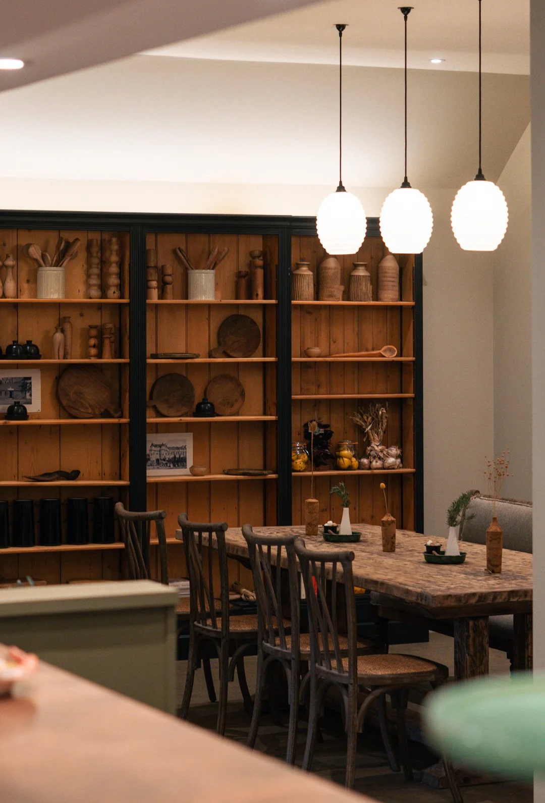 Wooden dining table with vases and decorative items, surrounded by wood chairs, against a wooden shelving unit with decorative objects and jars in a cozy interior setting.