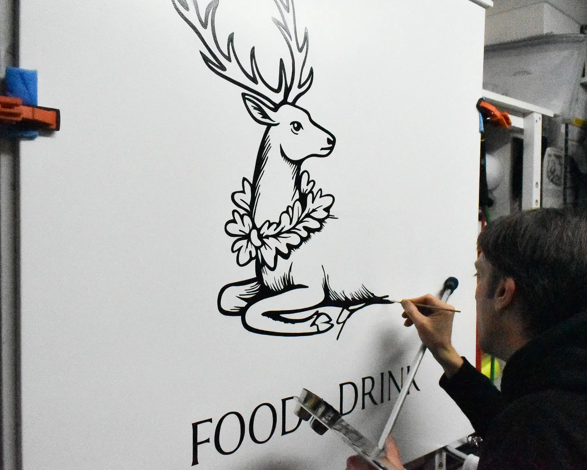 An artist painting a black and white logo of a reindeer's head with antlers, surrounded by a wreath, on a white board. Below the logo, the words 'FOOD & DRINK' are visible.