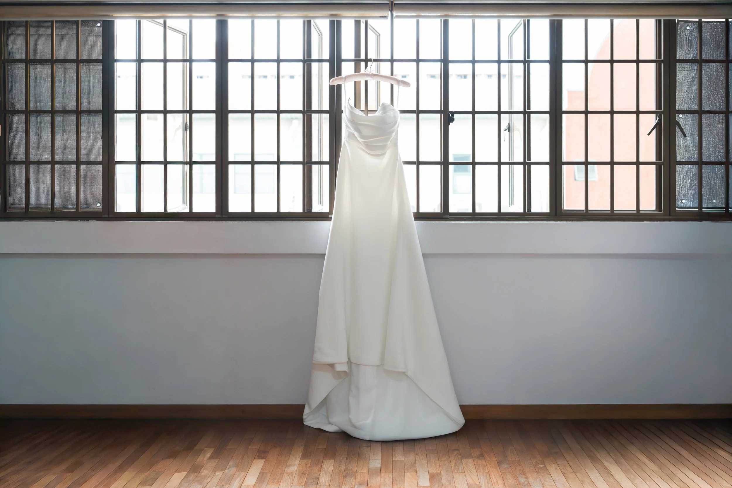 A white wedding dress hanging on a hanger in front of large multi-pane windows, with natural light illuminating the dress and the wooden floor below.