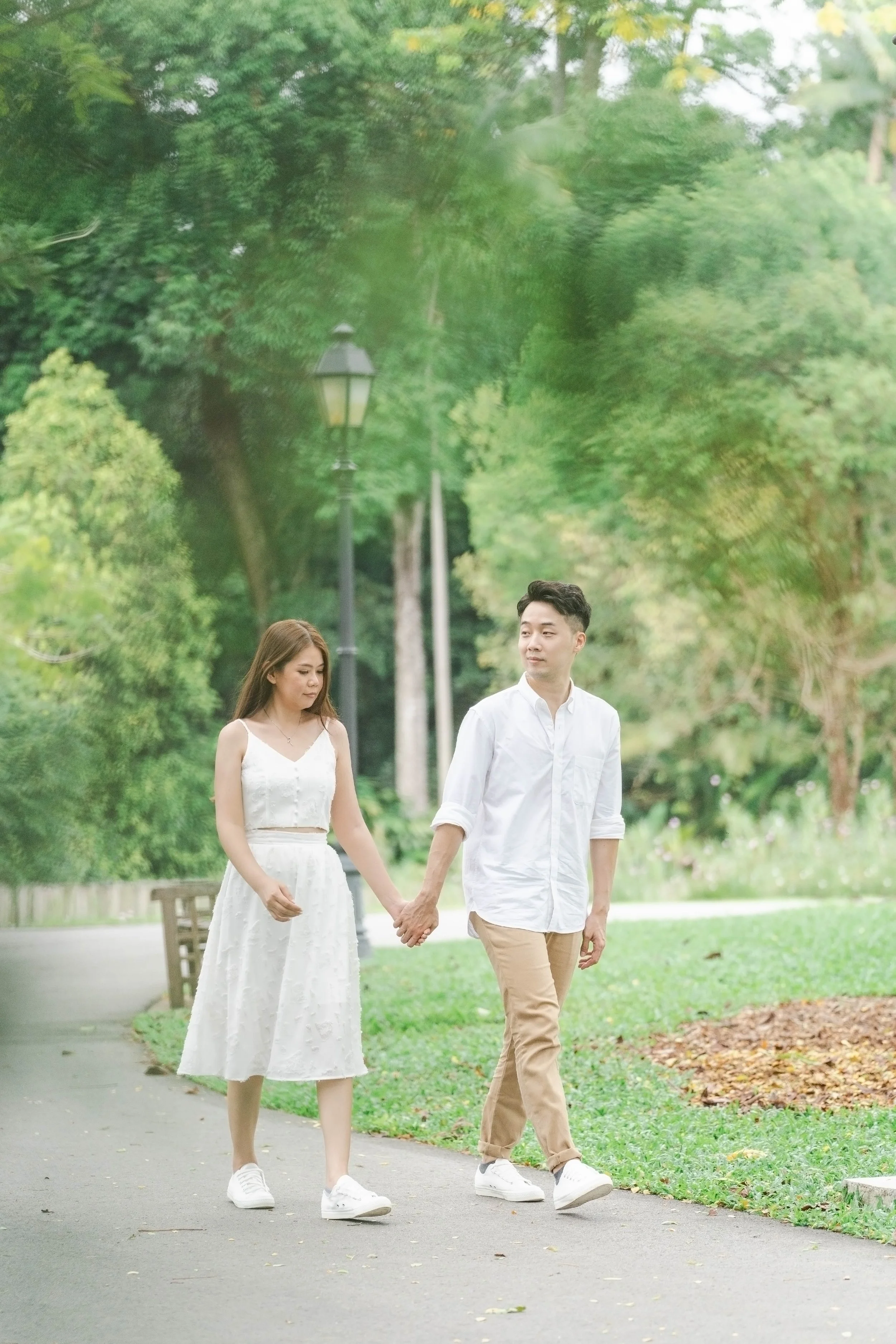 A young couple holding hands and walking in a park surrounded by green trees and grass.