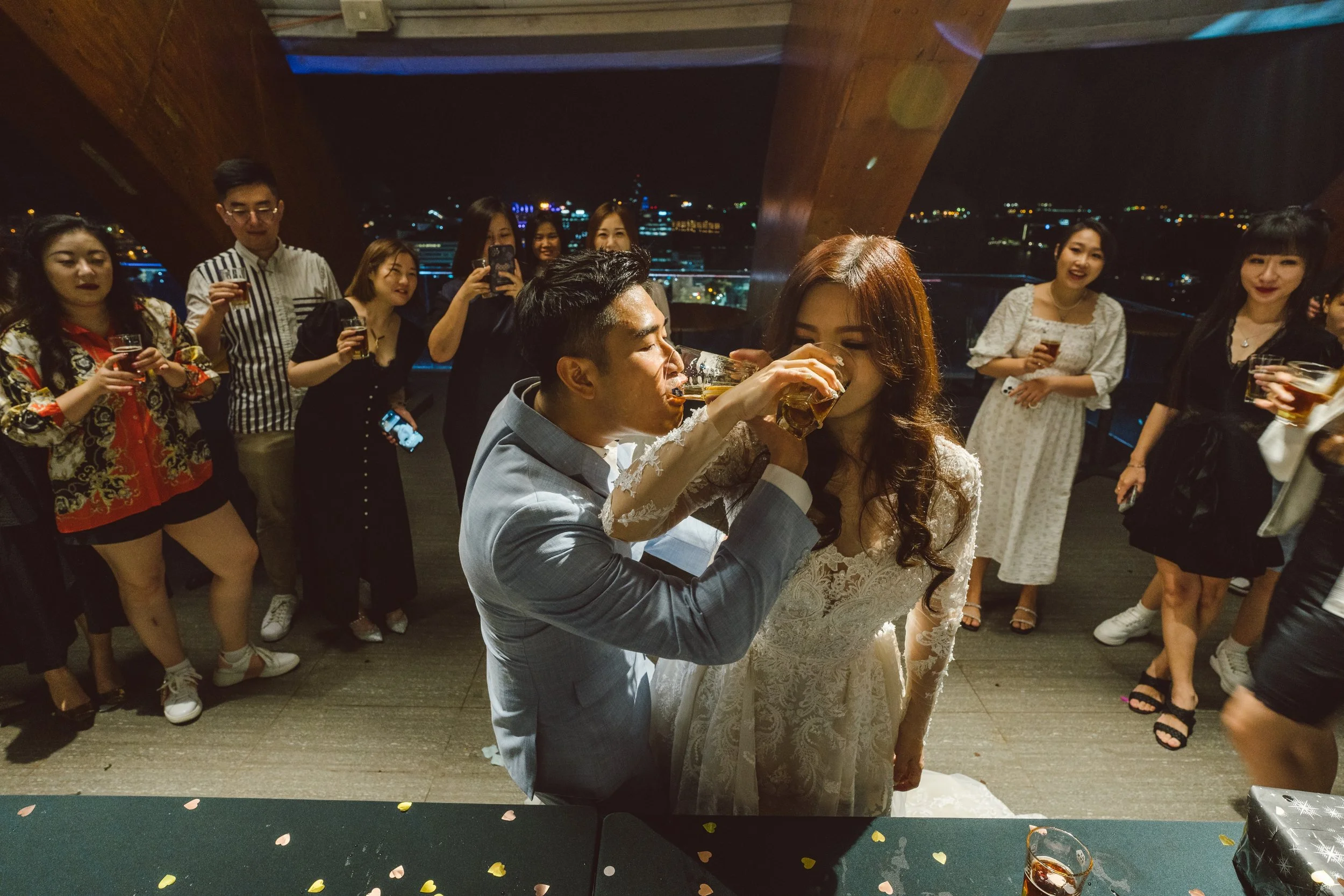 Korean wedding reception with bride and groom toasting drinks, surrounded by friends taking photos at night with citycity lights in the background.