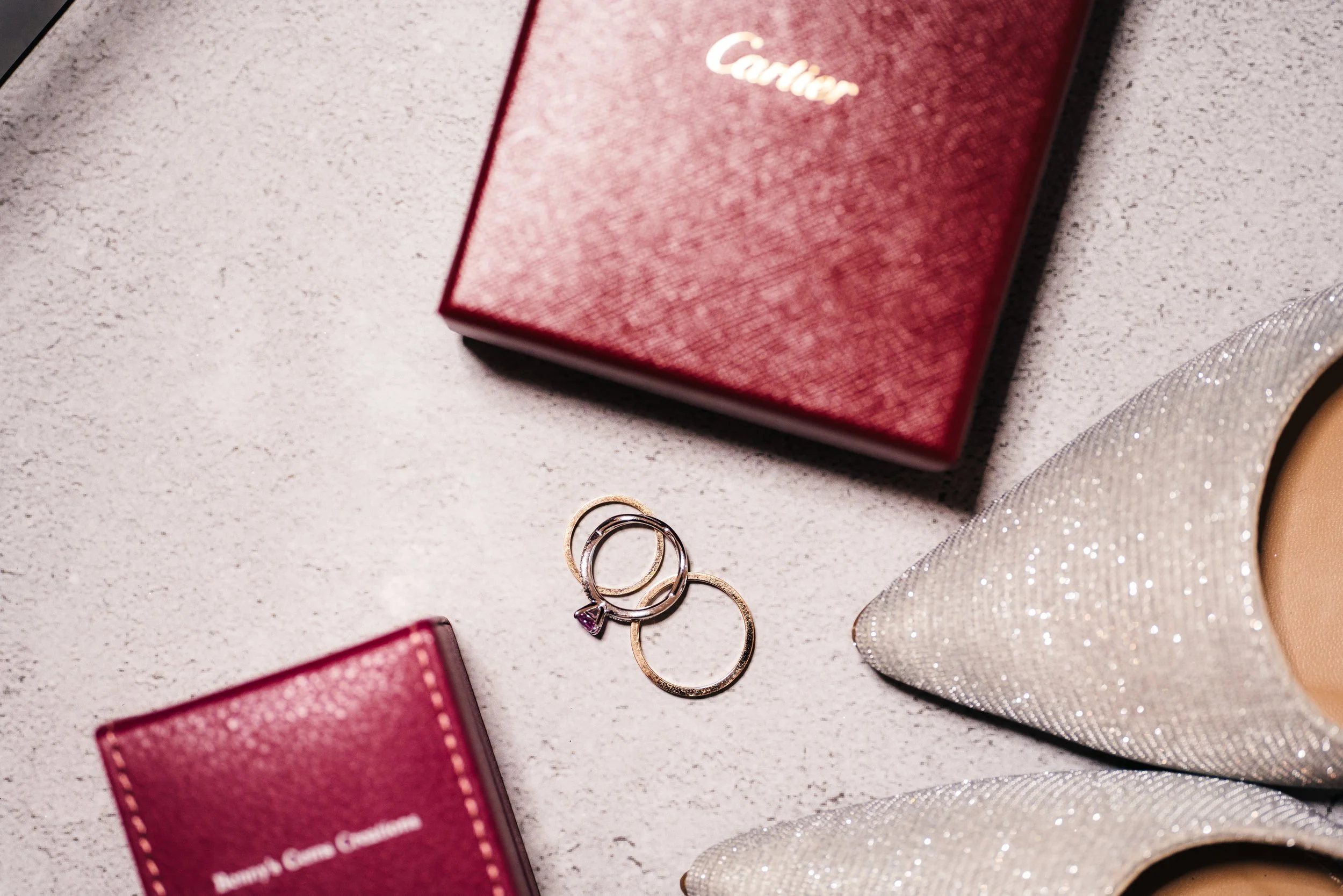 A pair of glittery high-heeled shoes, a red jewelry box with the word 'Camear' on it, and three rings, one with a purple stone, on a light-colored surface.