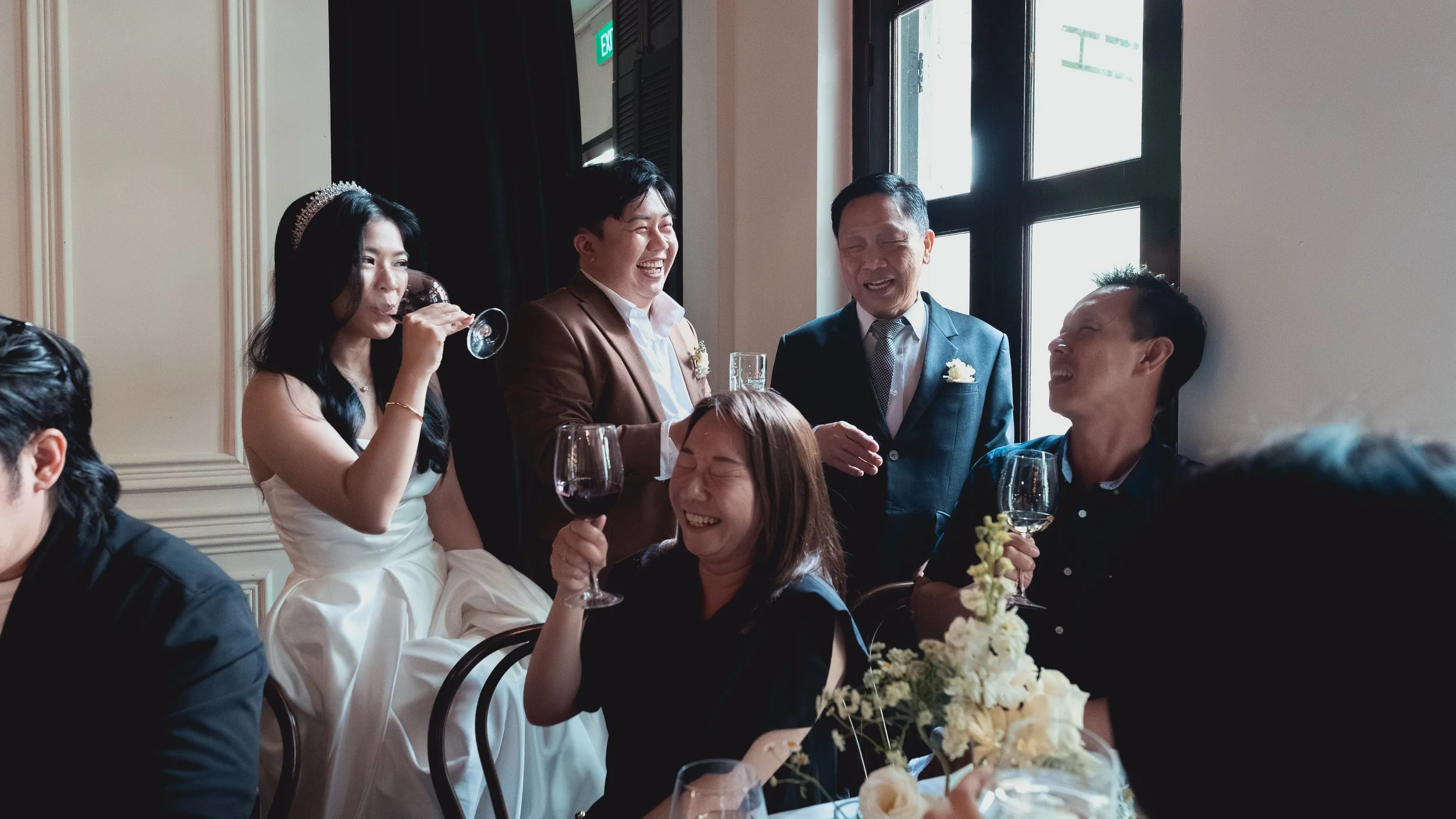 Group of six people at a celebration, with three standing and three sitting, laughing, holding glasses of red and white wine, near floral decorations and a window.