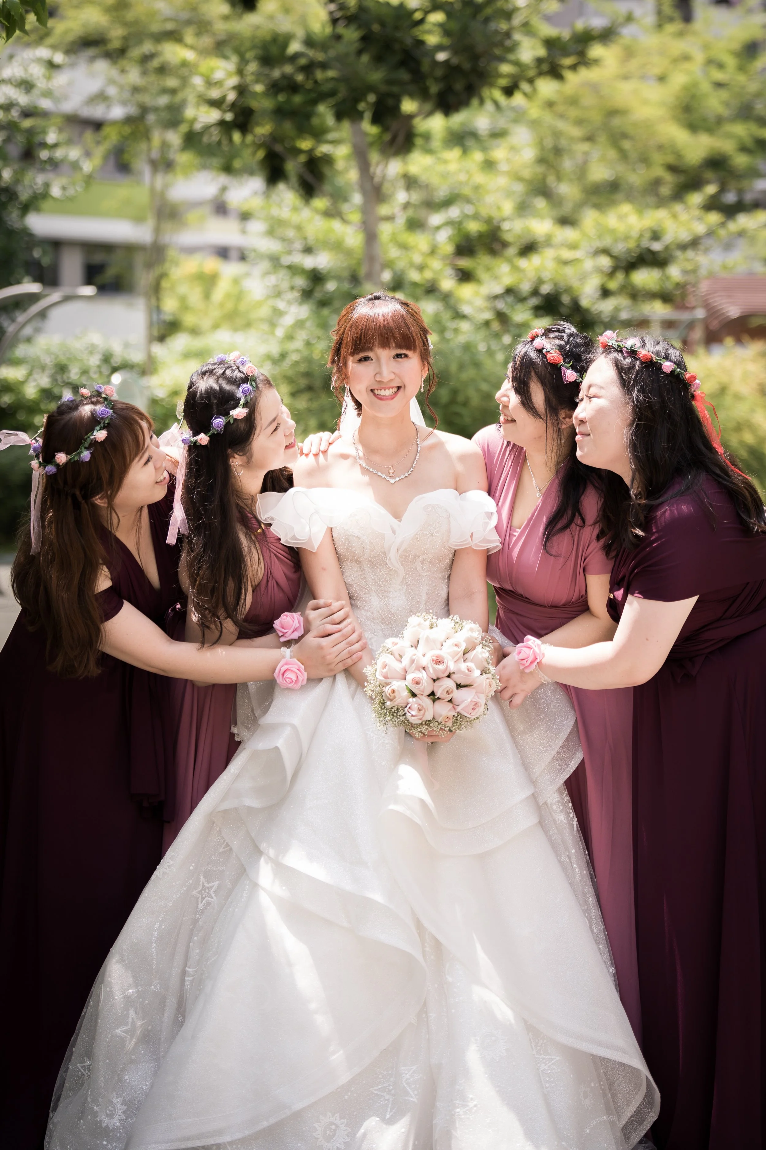 A bride in a white wedding gown holding a bouquet of pink roses is surrounded by five women in matching mauve dresses, all wearing flower crowns and pink wrist corsages, outdoors on a sunny day with trees in the background.