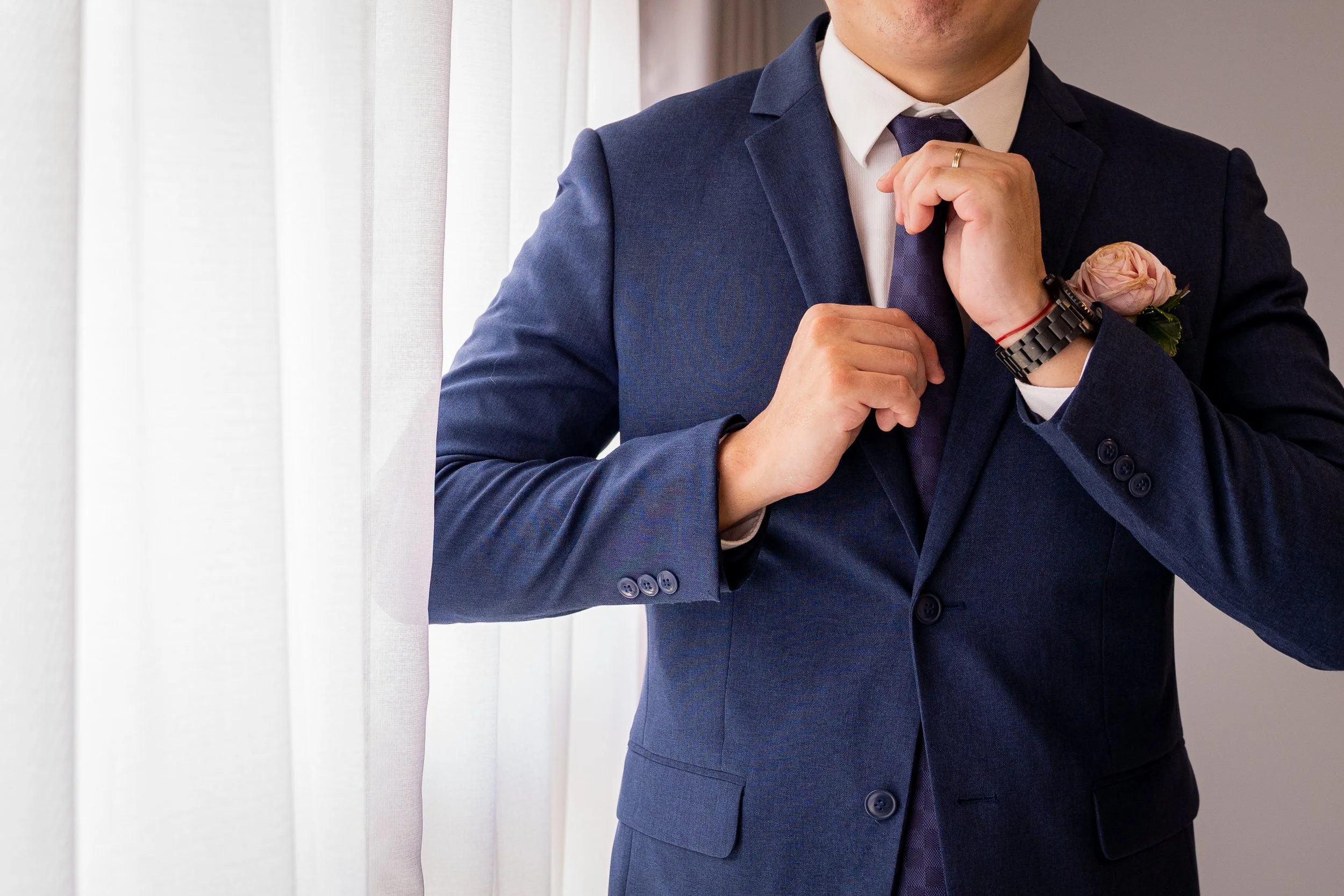 A man in a navy blue suit adjusting his tie, standing near white curtains, with a pink rose boutonniere on his lapel.