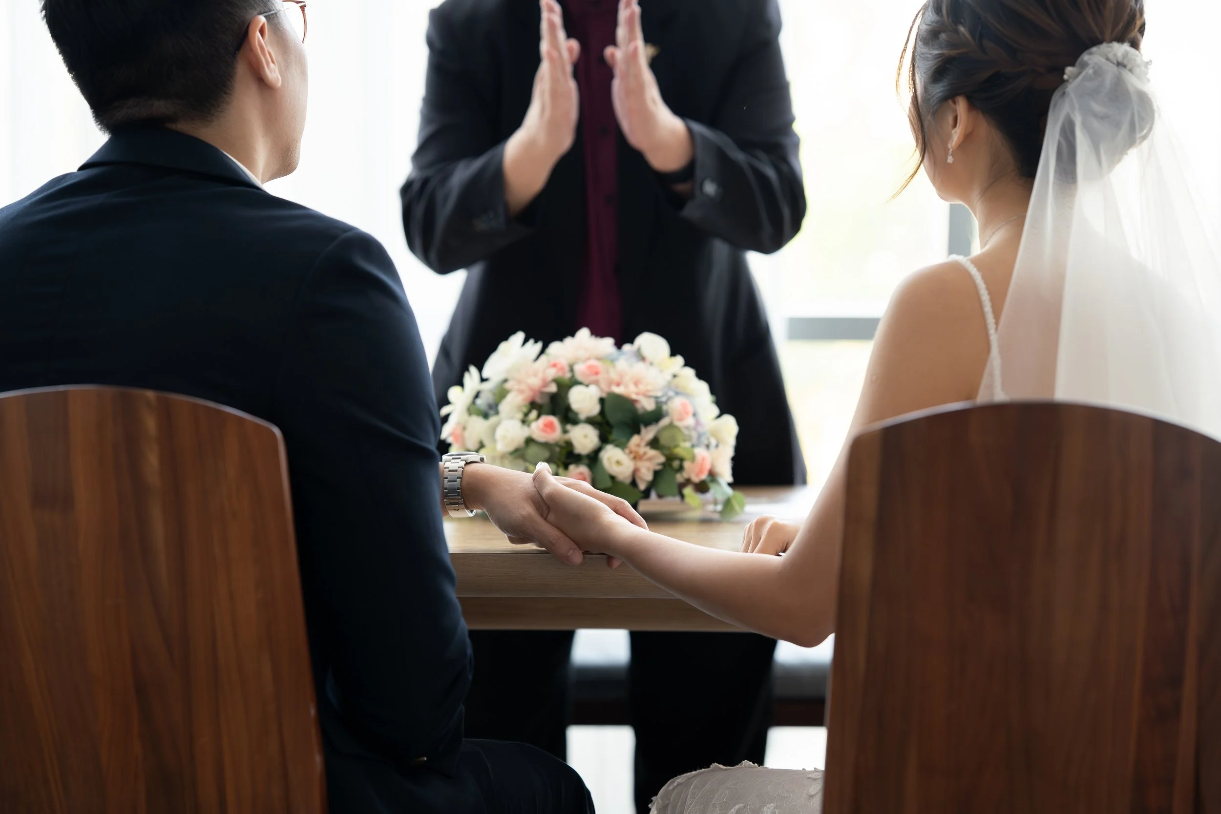 A bride and groom holding hands during a wedding ceremony, with an officiant standing behind them and a bouquet of flowers on the table.