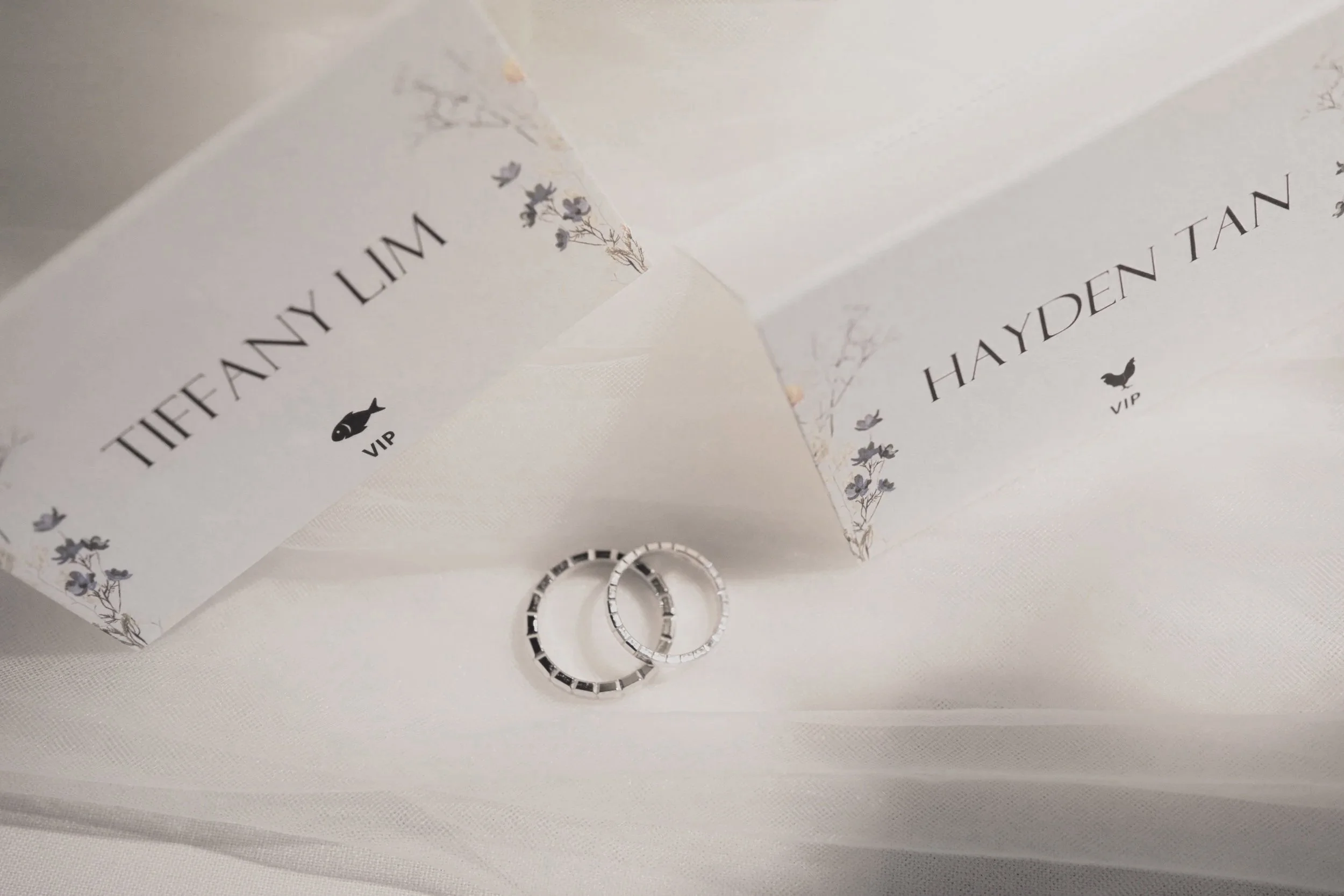 Two wedding rings placed on a light fabric, with wedding invitations behind them. The invitations display the names Tiffany Lim and Hayden Tan, with small floral illustrations and the word VIP.