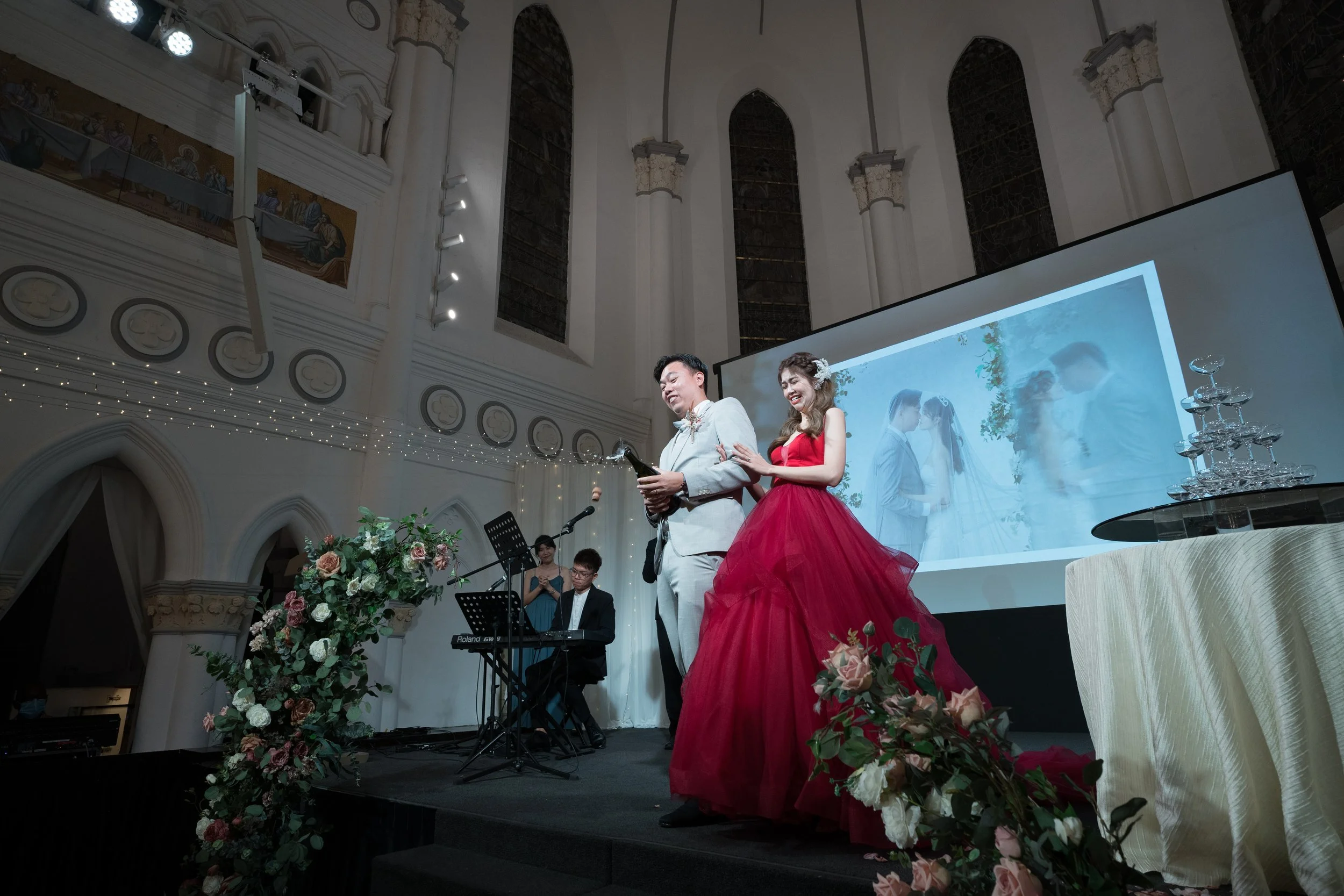A wedding ceremony with a bride and groom standing on stage, the bride in a red gown and the groom in a light-colored suit, with a music band playing nearby and a large screen projecting their wedding photo in the background, decorated with flowers a