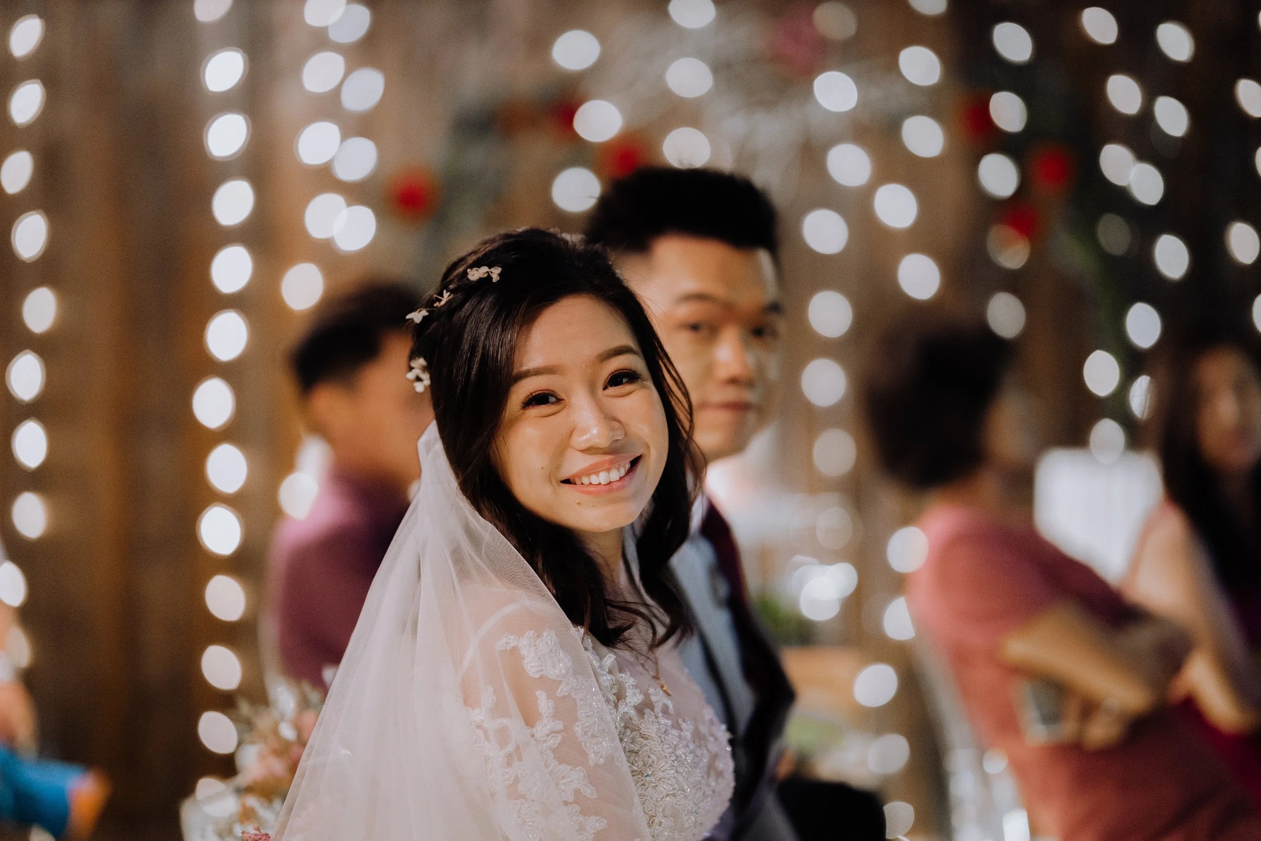 Happy bride in wedding dress and veil smiling at camera during wedding ceremony, with groom and guests in background, decorated with string lights.