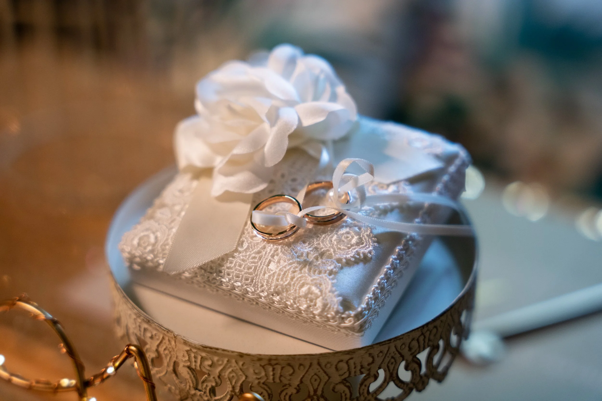 Close-up of a wedding ring set on a lace pillow, adorned with a ribbon and flower, placed inside an ornate metal dish.