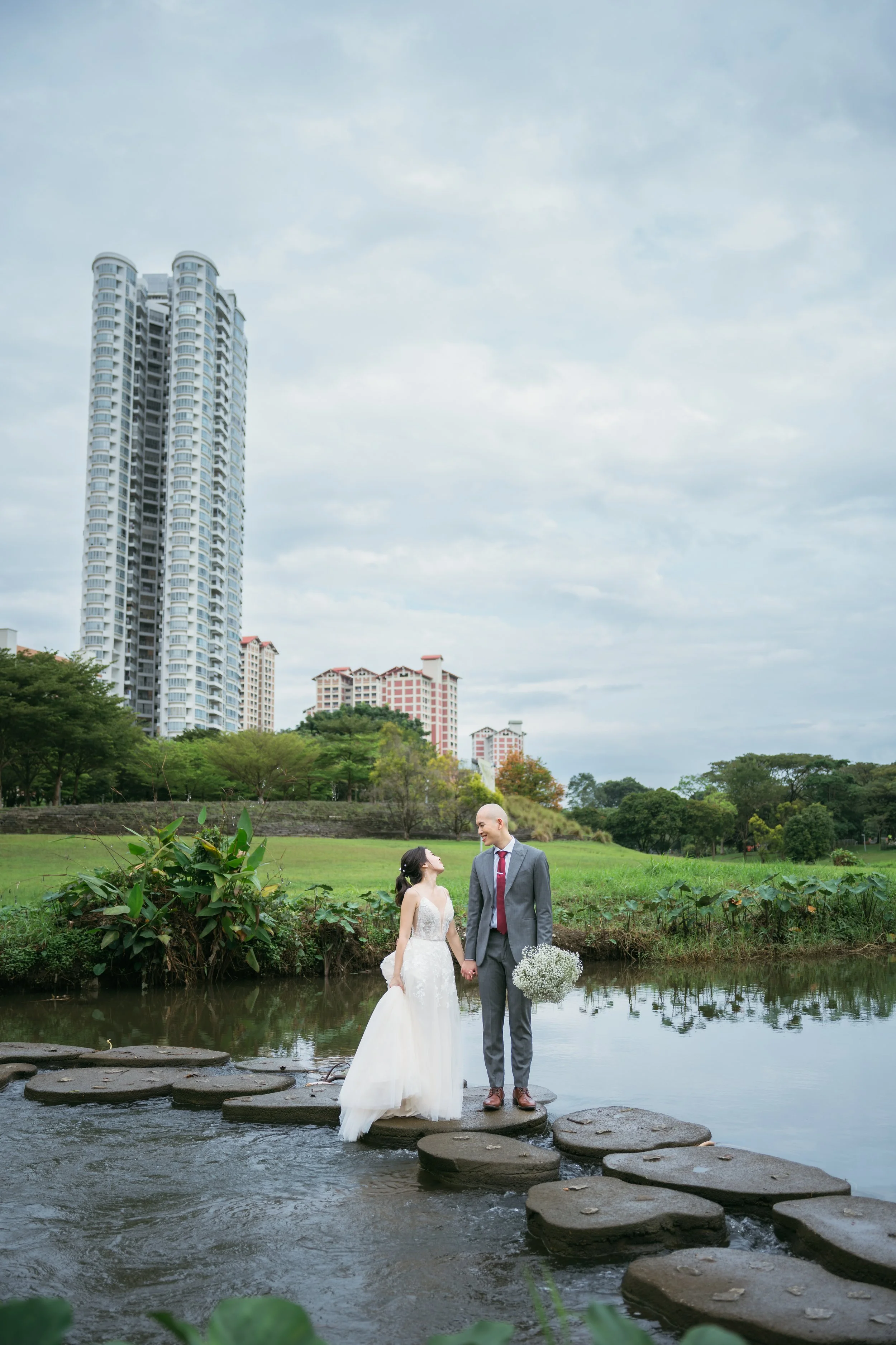A bride and groom holding hands and standing on stone steps in a river, with city's tall buildings and lush greenery in the background.